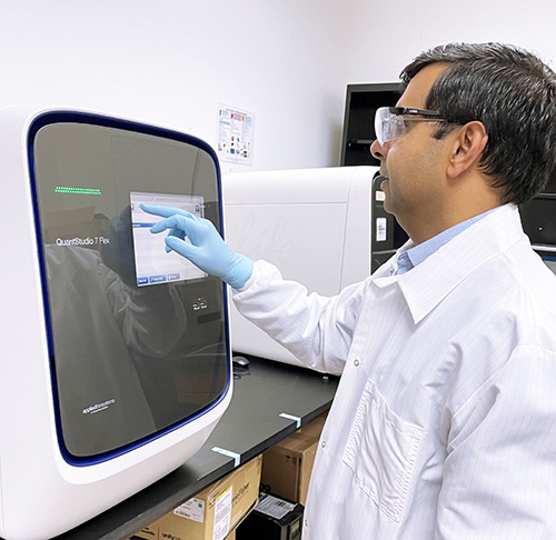 Technicien de laboratoire, en blouse blanche et lunettes de protection, en train d’utiliser l’appareil QuantStudio 7 Flex pour réaliser une analyse qPCR. Le technicien interagit avec l’écran tactile de l’instrument dans un environnement de laboratoire pharmaceutique moderne, dédié aux tests moléculaires, à la quantification d’ADN et à la recherche biomédicale. L’image illustre les capacités technologiques avancées d’Eurofins dans le domaine de la biologie moléculaire et du développement de médicaments.