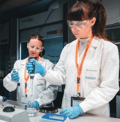 Deux techniciennes de laboratoire, équipés de blouses blanches, gants bleus et lunettes de protection, réalisent des manipulations précises à l’aide de pipettes dans un environnement de laboratoire Eurofins. Leurs badges arborent le logo Eurofins, soulignant leur appartenance à l’équipe. En arrière-plan, des équipements scientifiques spécialisés, dont un appareil marqué "TYPHOON", témoignent de l’environnement technologique avancé dédié aux essais analytiques dans le domaine pharmaceutique.