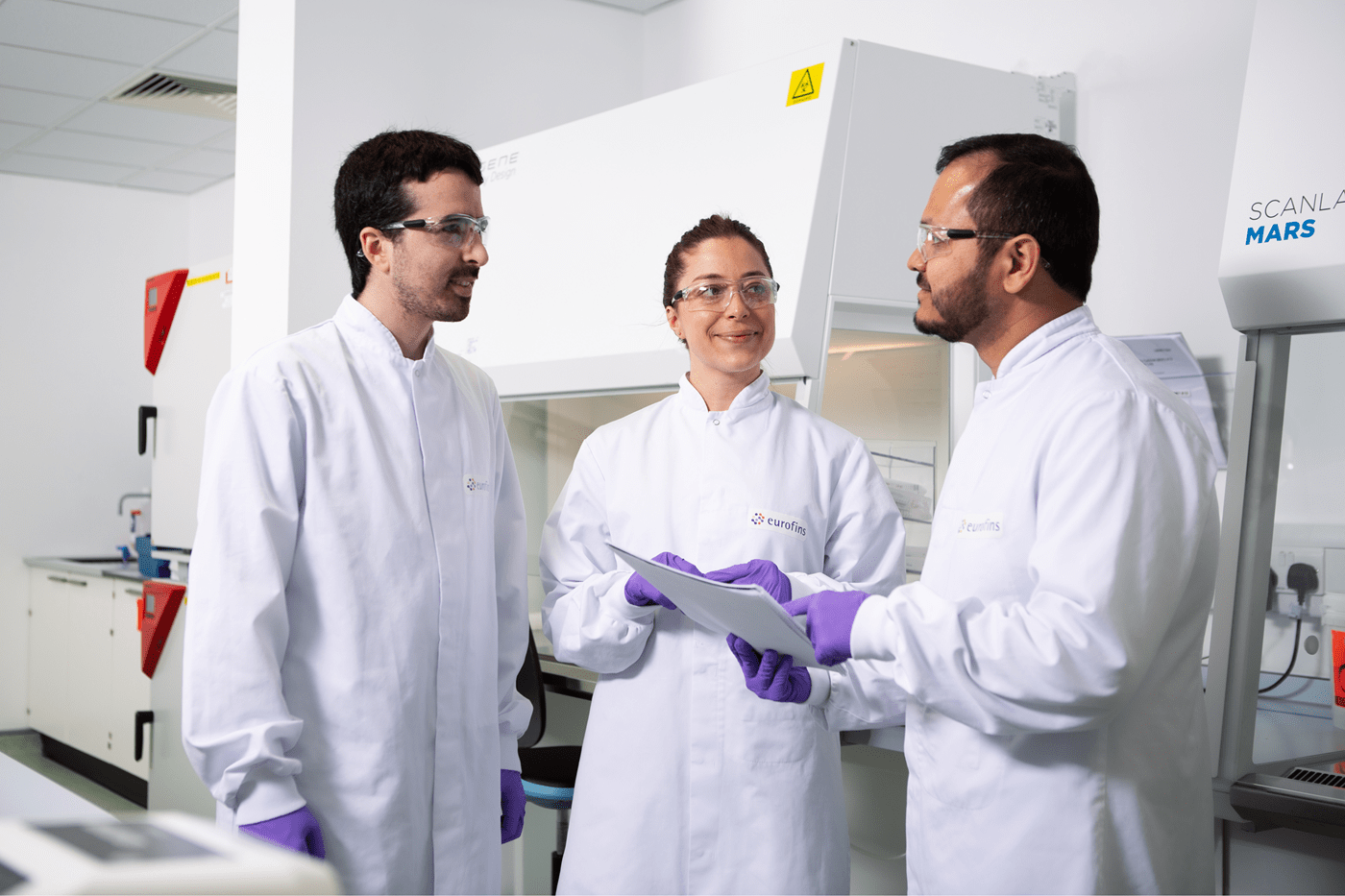 Trois membres de l’équipe Eurofins Biopharma Product Testing discutant dans un laboratoire devant une hotte de sécurité. Ils portent des blouses Eurofins et des gants de protection, avec un document à la main, dans un environnement scientifique moderne équipé pour les analyses pharmaceutiques.