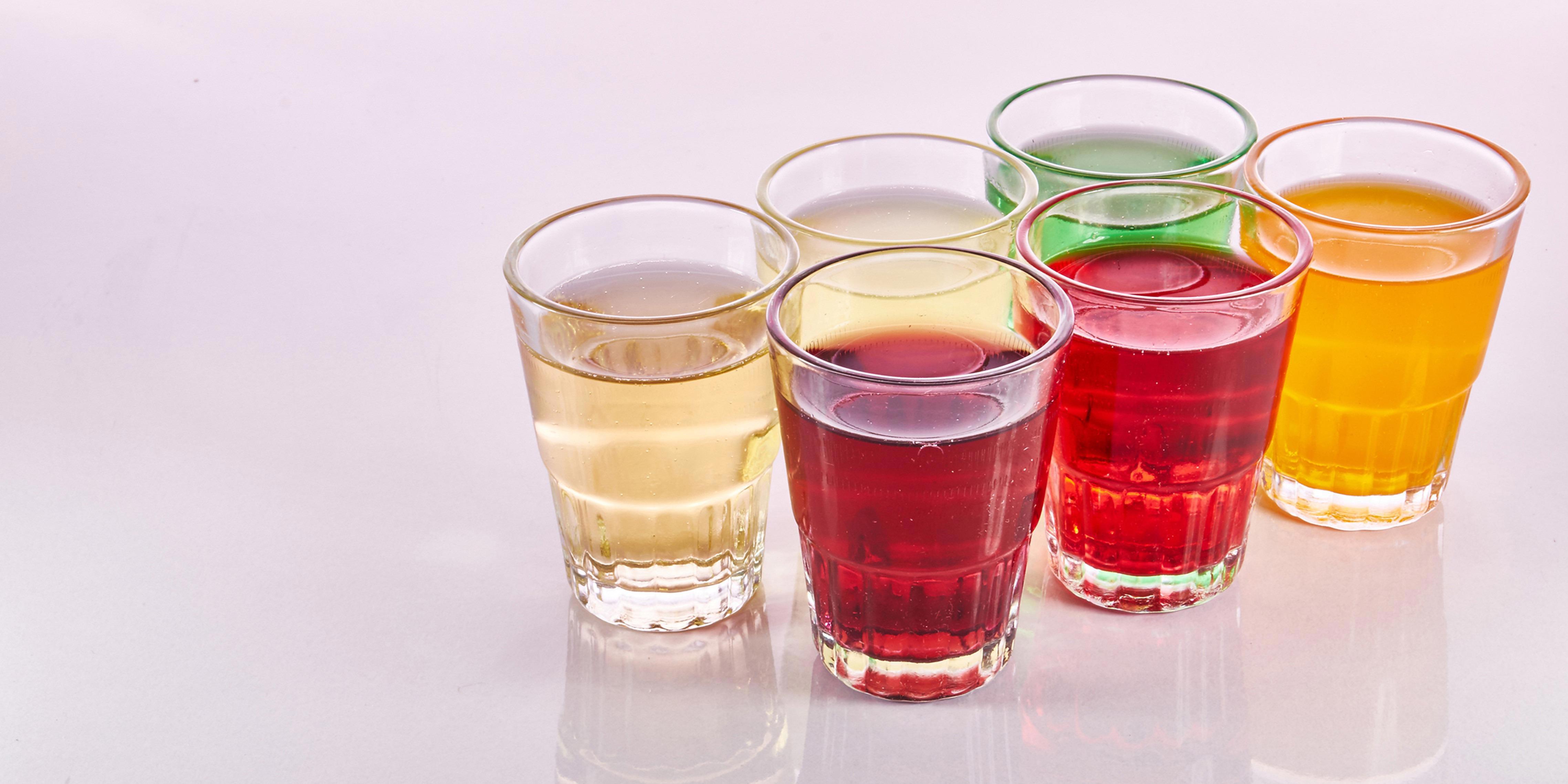 Six small glasses filled with different coloured liquids are arranged in two diagonal rows on a reflective surface. The liquids are yellow, red, green, and orange. The reflection of the glasses and their contents is visible on the surface below them.