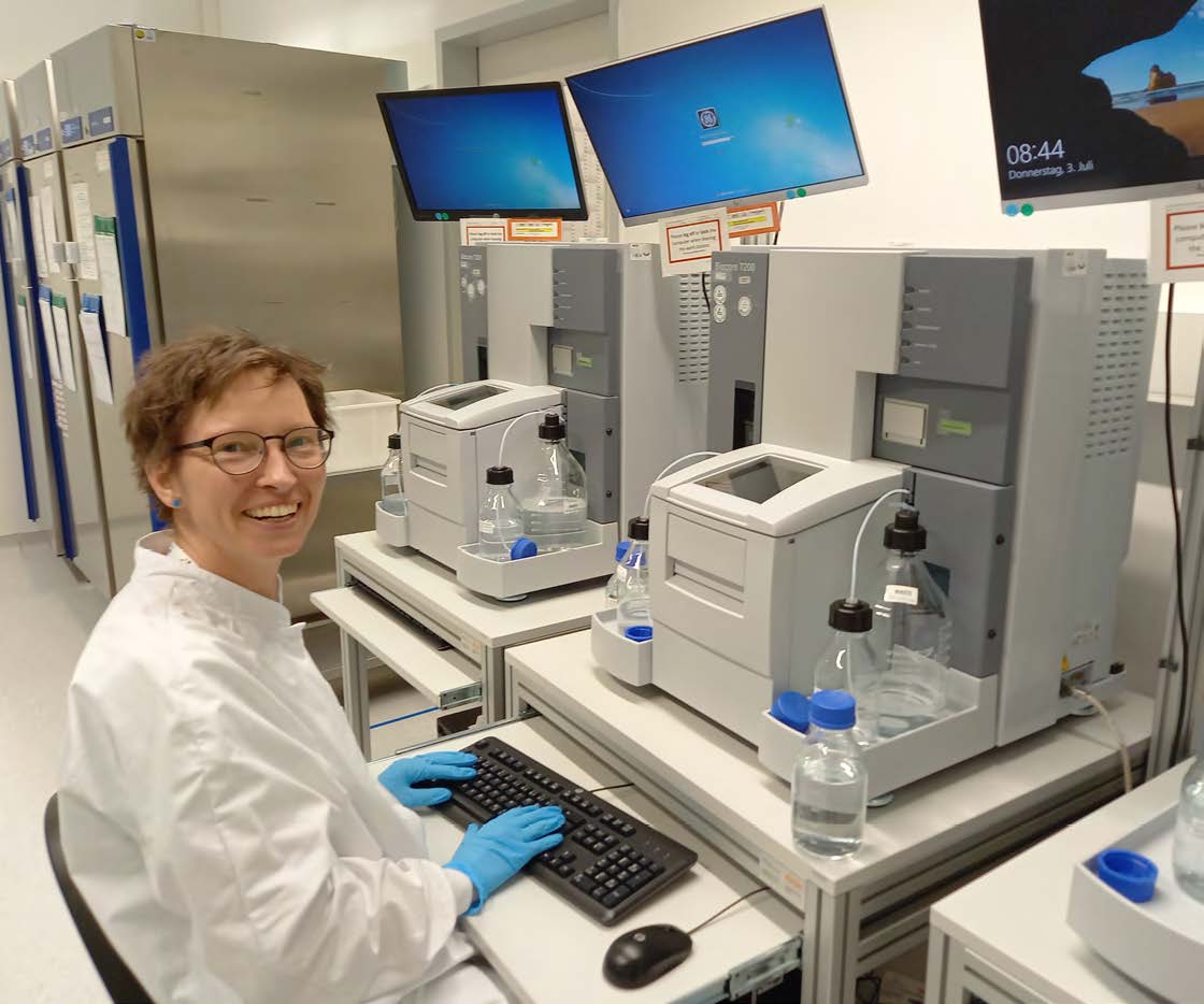 Personne en blouse de laboratoire et gants bleus assise devant un ordinateur dans un environnement scientifique. En arrière-plan, plusieurs équipements de laboratoire, flacons et machines sont visibles, accompagnés de deux écrans d’ordinateur. La personne regarde vers la caméra avec un sourire. Cette scène illustre les activités de laboratoire menées par Eurofins BioPharma Product Testing France.