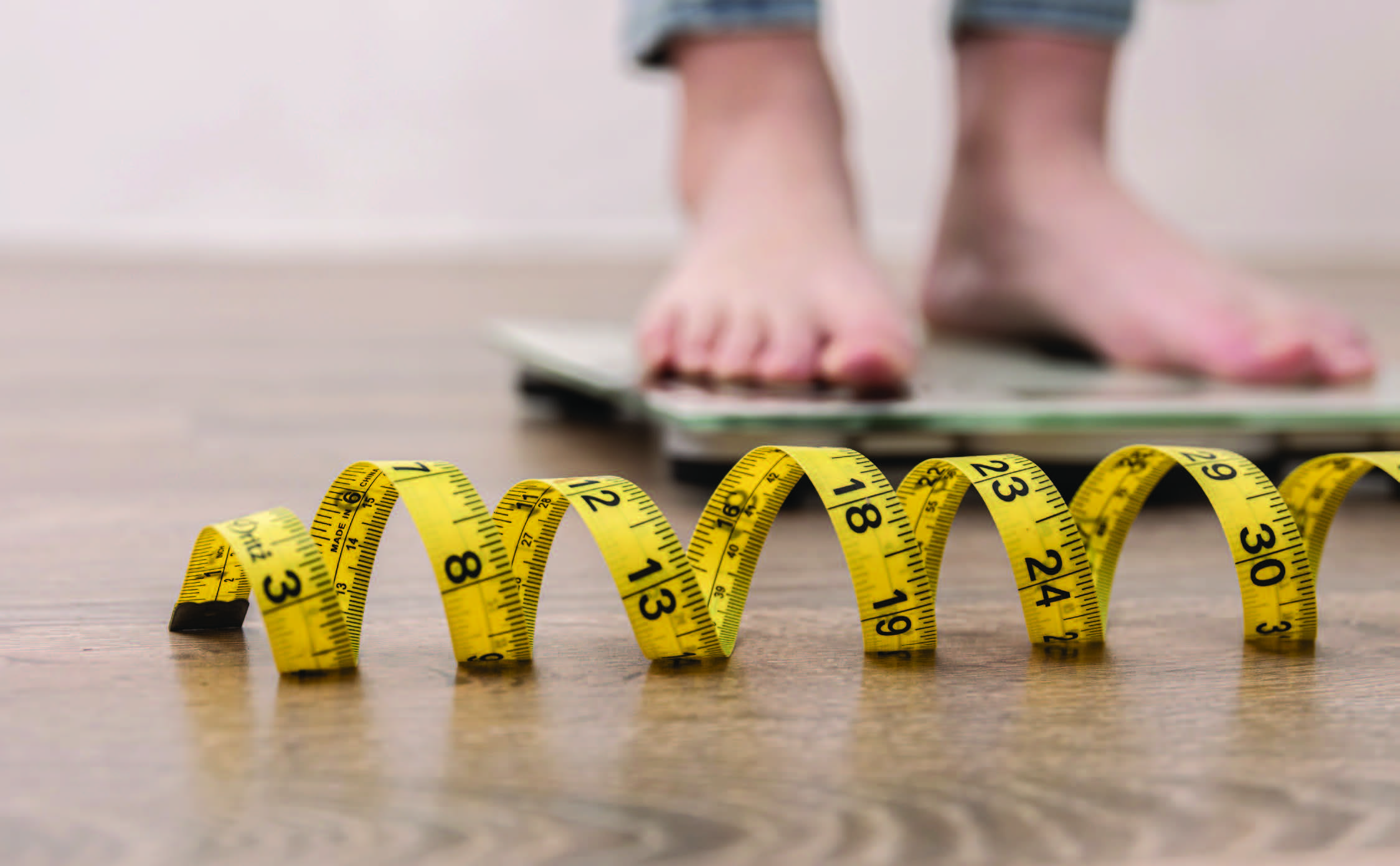 Gros plan sur un mètre ruban jaune enroulé au sol, avec en arrière-plan une personne debout pieds nus sur une balance. L’image illustre des thématiques liées à la mesure du poids, à la santé ou au suivi de la condition physique, dans le cadre des activités ou des études menées par Eurofins BioPharma Product Testing France.