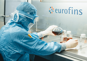 Technicien en tenue stérile manipulant des solutions sous hotte à flux laminaire dans un laboratoire Eurofins BioPharma Product Testing, réalisant des tests de stérilité et analyses microbiologiques pour le contrôle qualité des produits pharmaceutiques. Équipement de protection complet incluant gants, masque et combinaison, garantissant la conformité aux normes GMP et ISO pour la sécurité des patients et la fiabilité des résultats.