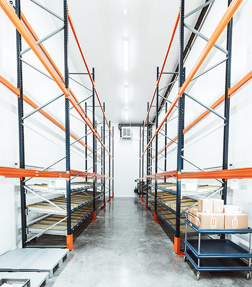 Zone de stockage Eurofins propre et bien éclairée, avec rayonnages métalliques hauts partiellement remplis de cartons. Un chariot bleu chargé de boîtes en carton est visible au premier plan. Le sol en béton poli et l’éclairage fluorescent renforcent l’aspect organisé et industriel de l’espace, utilisé pour le stockage de produits pharmaceutiques dans le cadre d’études de stabilité ou de contrôle qualit