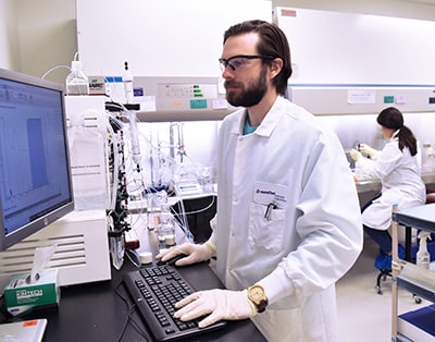 Technicien de laboratoire chez Eurofins BioPharma Product Testing travaillant sur un poste pour analyser des données expérimentales. Sur la paillasse, des instruments de chromatographie et des systèmes de tubulures sont utilisés pour la purification et la validation des échantillons. À l’arrière-plan, une hotte de sécurité biologique est employée pour des manipulations stériles, illustrant les standards élevés de qualité et de sécurité appliqués par Eurofins dans ses services analytiques.