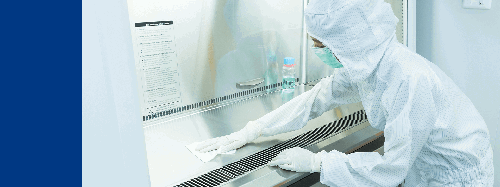 Opération de nettoyage dans une zone stérile chez Eurofins BioPharma Product Testing. Une personne porte un équipement complet de protection (combinaison intégrale, gants et masque) et désinfecte la surface d’une hotte à flux laminaire, illustrant les procédures strictes de décontamination et de conformité aux normes GMP pour garantir la sécurité des analyses biopharmaceutiques. 