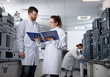 Laboratoire Eurofins BioPharma Product Testing dédié à l’identification et à la quantification de solvants ou d’impuretés inconnus. Deux personnes en blouse blanche consultent un classeur bleu au centre de la pièce, tandis qu’une autre travaille sur un instrument analytique. La salle est équipée de plusieurs systèmes de chromatographie et d’analyse de haute précision, illustrant les standards de qualité et de conformité appliqués aux tests biopharmaceutiques.