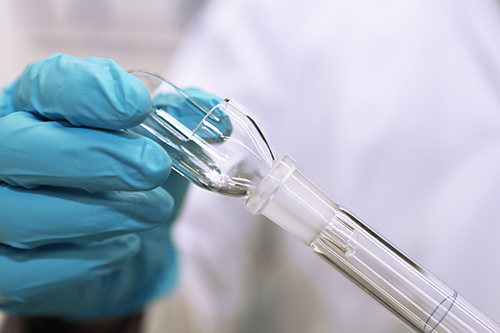 Main gantée de bleu manipulant un tube en verre dans un laboratoire Eurofins, insérant le tube dans un équipement en verre pour une préparation expérimentale. L’image met en avant la précision des gestes en environnement scientifique, illustrant les pratiques de manipulation de standards de référence dans le cadre de tests analytiques et de contrôle qualité pharmaceutique.
