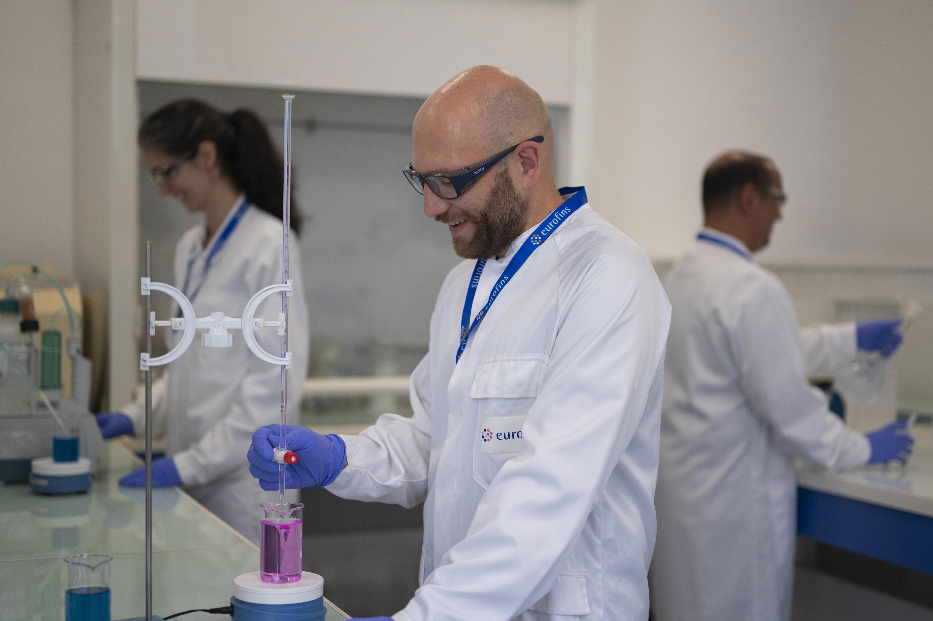 Techniciens de laboratoire réalisant des manipulations analytiques, dont une titration avec une burette contenant un liquide rose, illustrant l’engagement d’Eurofins BioPharma Product Testing face aux défis posés par les PFAS dans l’industrie pharmaceutique.