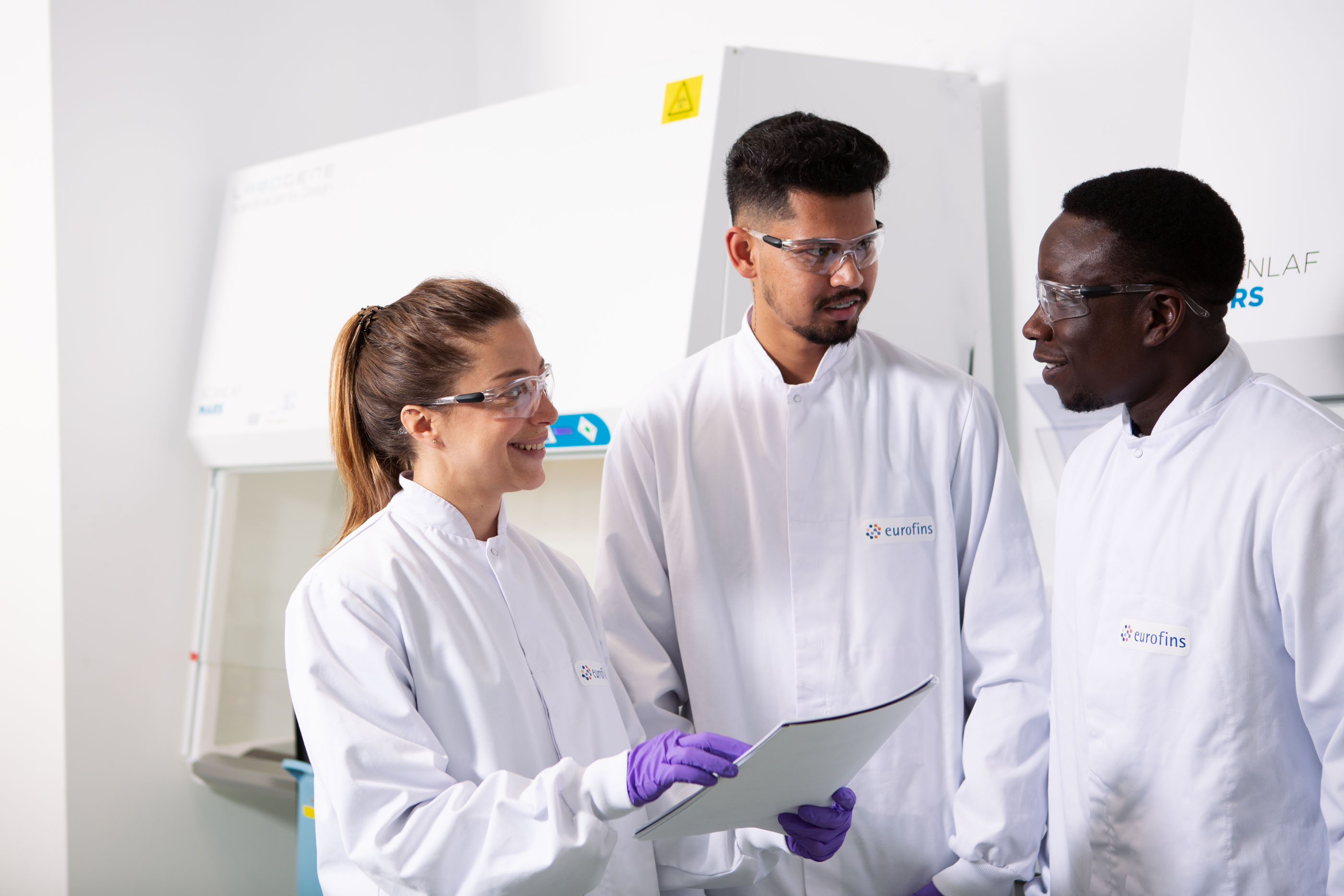 Trois scientifiques Eurofins en blouse blanche, équipés de lunettes de protection, discutent dans un laboratoire pharmaceutique devant une enceinte de sécurité biologique, illustrant la collaboration, la rigueur scientifique et les protocoles de sécurité appliqués lors des études de stabilité et des tests de libération de médicaments et de substances actives.