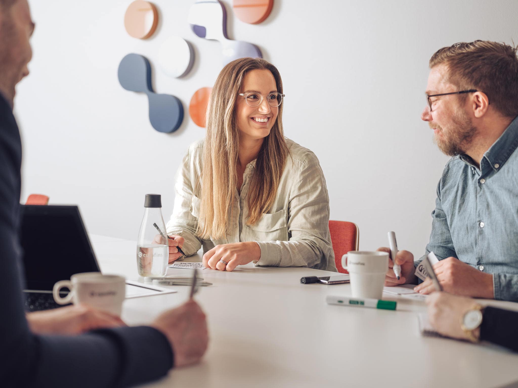 Réunion d’équipe Eurofins BioPharma Product Testing autour des services harmonisés : des collaborateurs échangent dans une salle moderne, illustrant la coordination dans les projets pharmaceutiques. Un ordinateur portable, des documents et des tasses de café sont disposés sur la table, renforçant l’ambiance collaborative. La décoration murale colorée en arrière-plan reflète l’engagement d’Eurofins pour l’innovation, la qualité et l’harmonisation des processus analytiques et réglementaires.