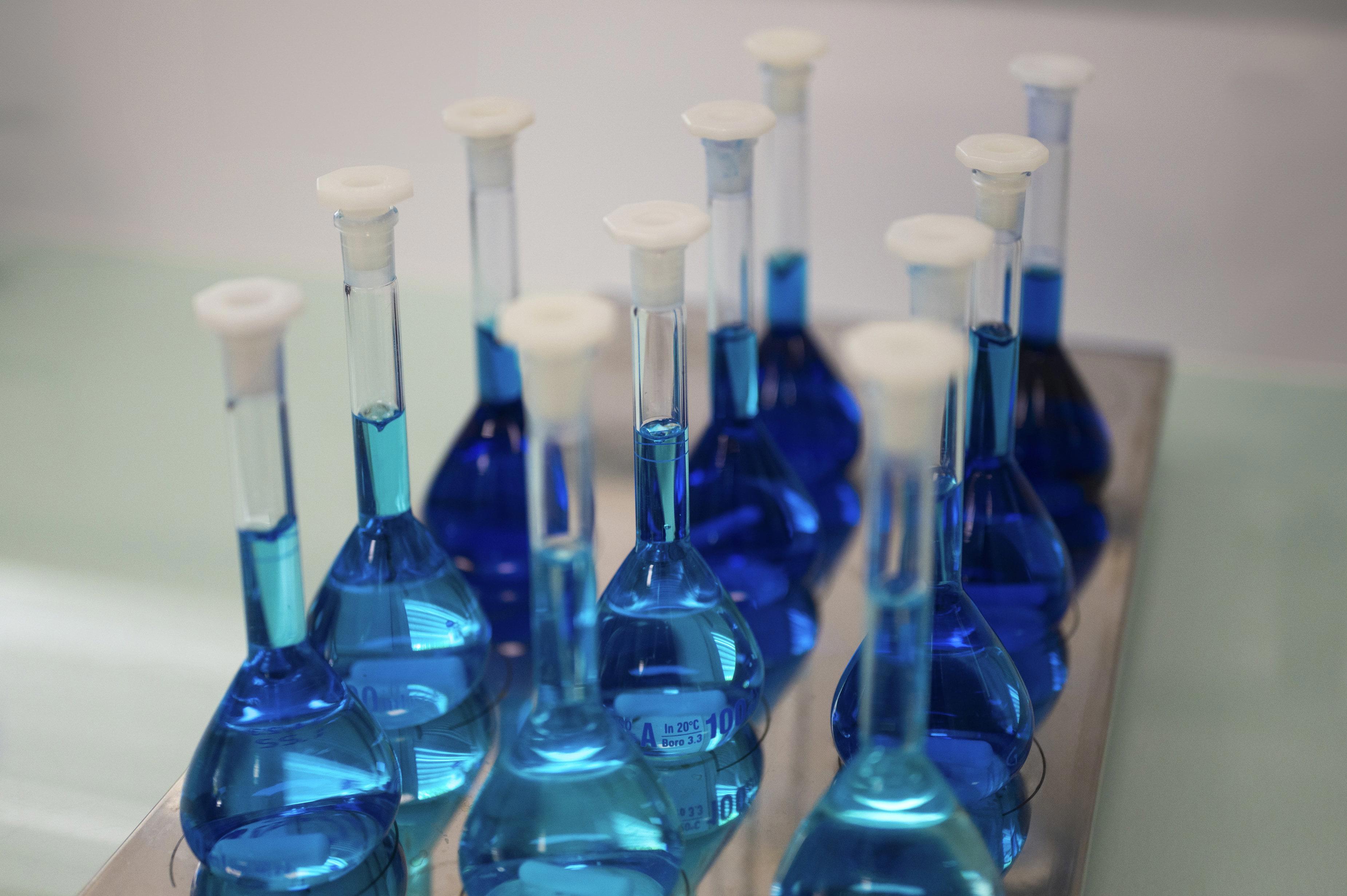 Plusieurs fioles jaugées en verre transparent, remplies d’une solution bleue, disposées sur un plateau métallique dans un environnement de laboratoire. Chaque fiole est fermée par un bouchon blanc. L’image illustre des préparations chimiques ou analytiques réalisées dans le cadre de tests en laboratoire, en lien avec les activités d’Eurofins BioPharma Product Testing France.