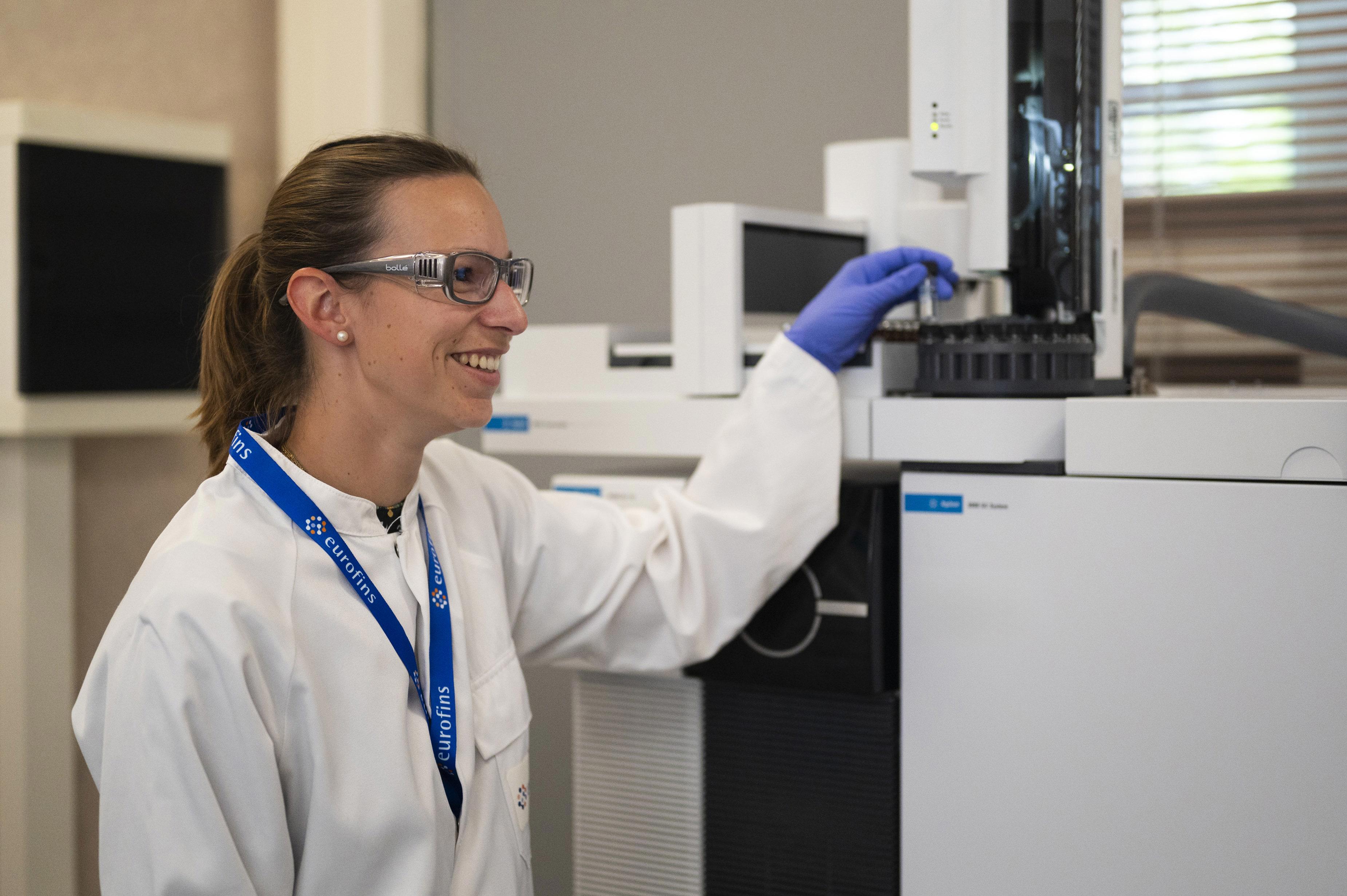 Technicienne de laboratoire souriant, équipé de lunettes de protection, blouse blanche et gants bleus, manipulant un instrument scientifique dans un environnement de laboratoire moderne. Illustration du professionnalisme et des protocoles de sécurité appliqués chez Eurofins Biopharma Product Testing.