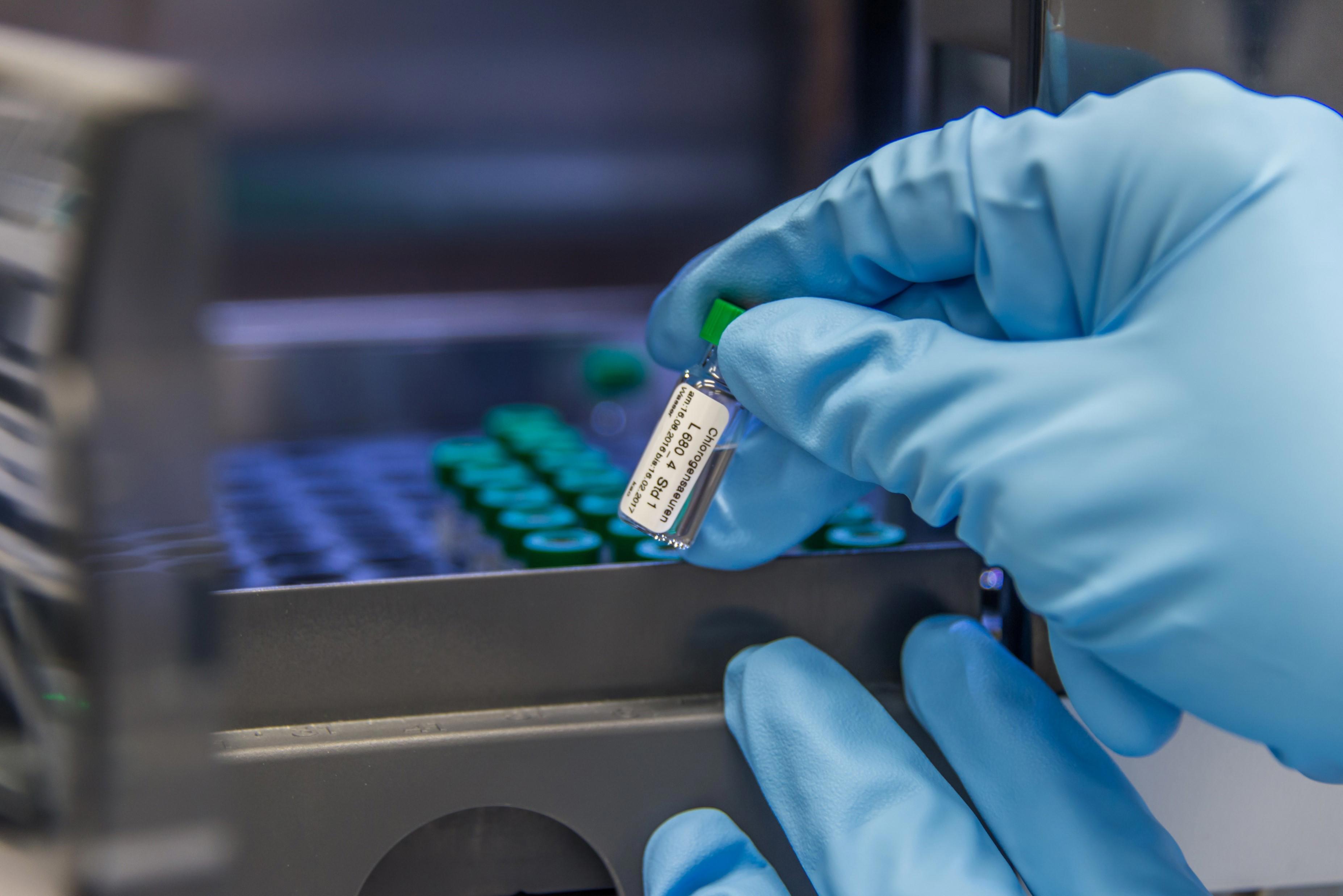 A close-up image of a gloved hand holding a small vial inside a laboratory setting. The background shows multiple similar vials arranged in rows within a storage unit or machine. 