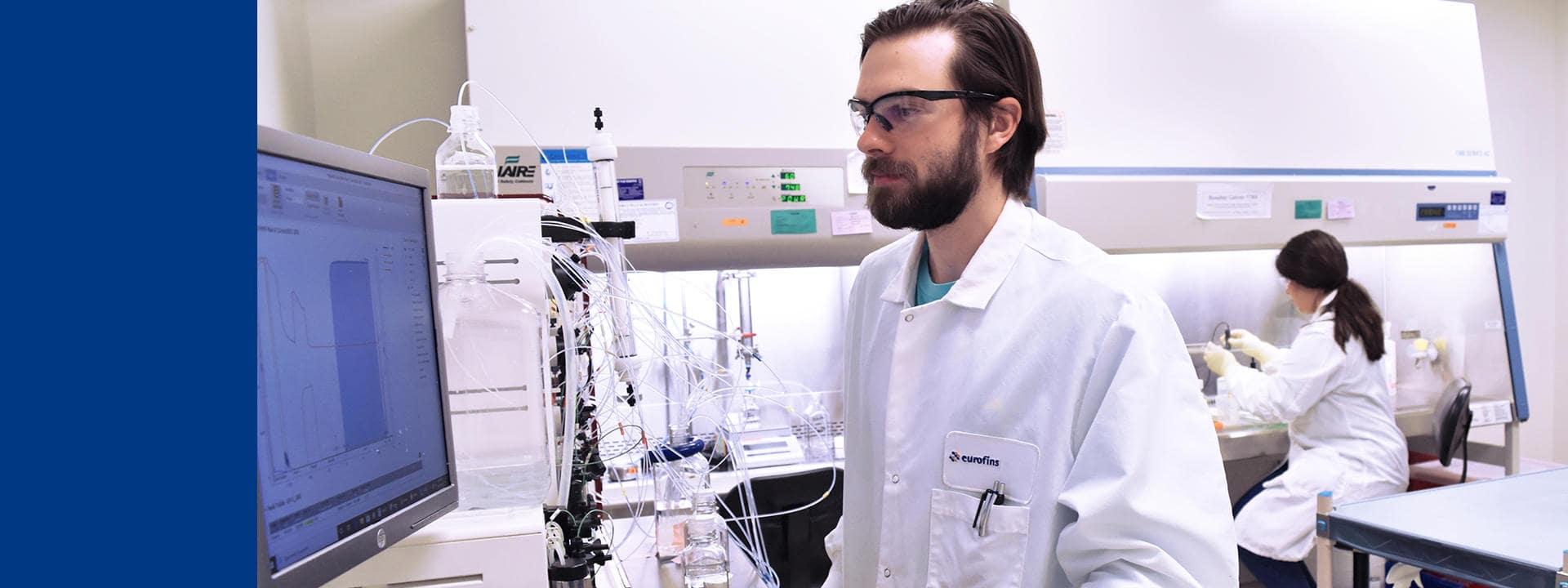 Laboratoire équipé pour des analyses biopharmaceutiques avancées. Au premier plan, un poste de travail avec un écran affichant des données expérimentales et des instruments de chromatographie reliés par des tubulures pour la purification et le contrôle qualité des échantillons. À l’arrière-plan, une zone sous hotte de sécurité biologique où des opérations de manipulation stérile sont réalisées par Eurofins BioPharma Product Testingdans ses services analytiques.