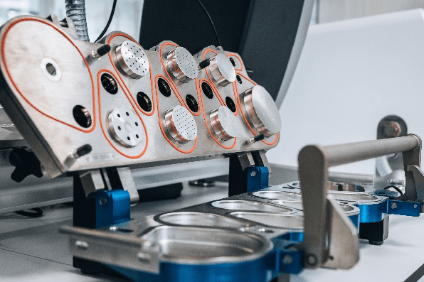 Équipement de laboratoire utilisé par Eurofins BioPharma Product Testing, composé de plusieurs compartiments circulaires à couvercles perforés et poignées bleues métalliques. Chaque compartiment est intégré dans une structure métallique avec des contours rouges, suggérant une utilisation pour la préparation ou l’analyse simultanée d’échantillons, notamment dans le cadre de tests d’inhalation ou de nébulisation.