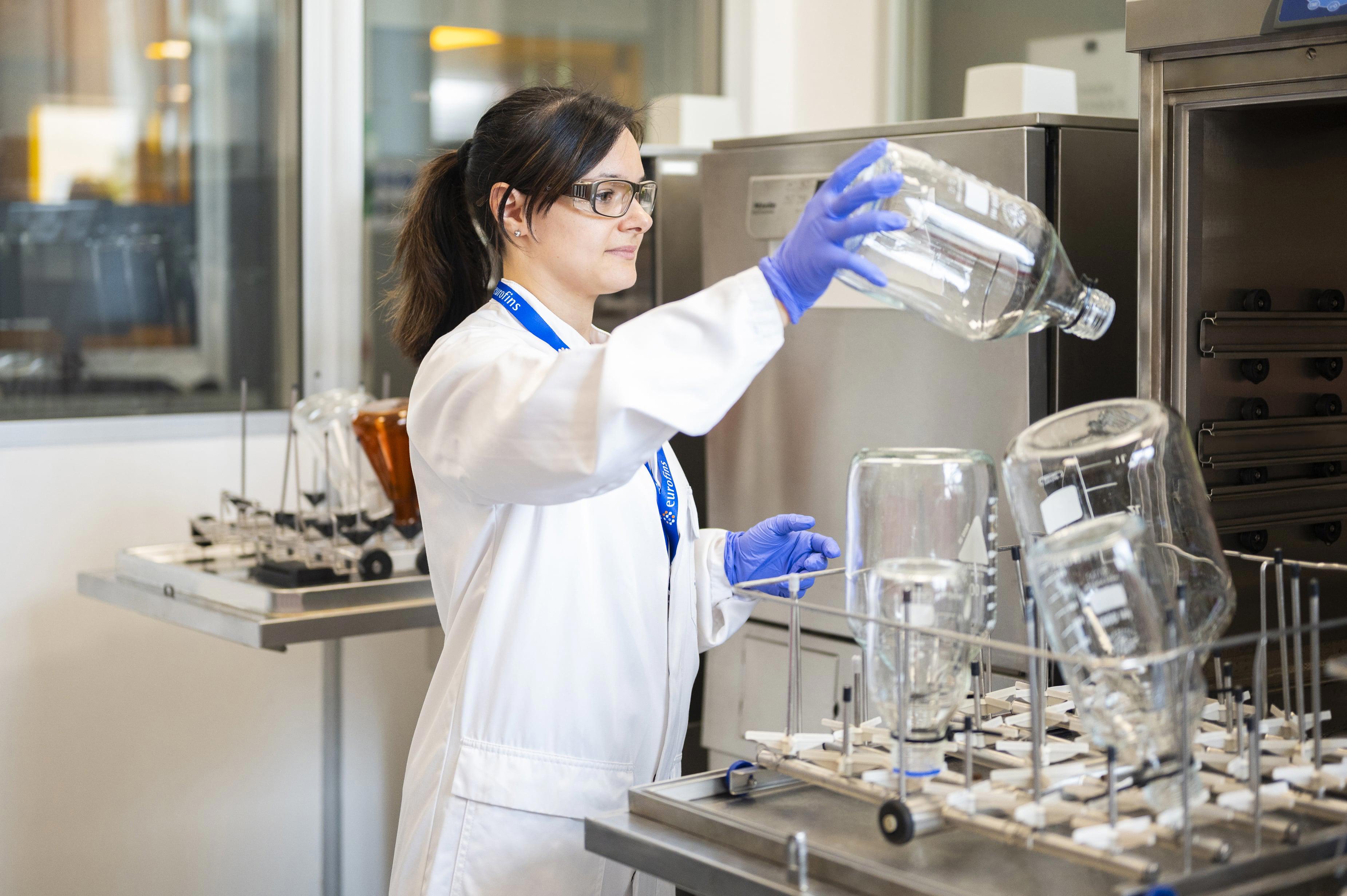 Technicienne de laboratoire portant une blouse blanche et des gants violets, manipulant de grands flacons en verre dans un environnement scientifique. L’image illustre les opérations de nettoyage et de préparation des équipements réalisées par Eurofins BioPharma Product Testing pour garantir l’efficacité du cycle de lavage et la conformité aux normes d’hygiène pharmaceutique.