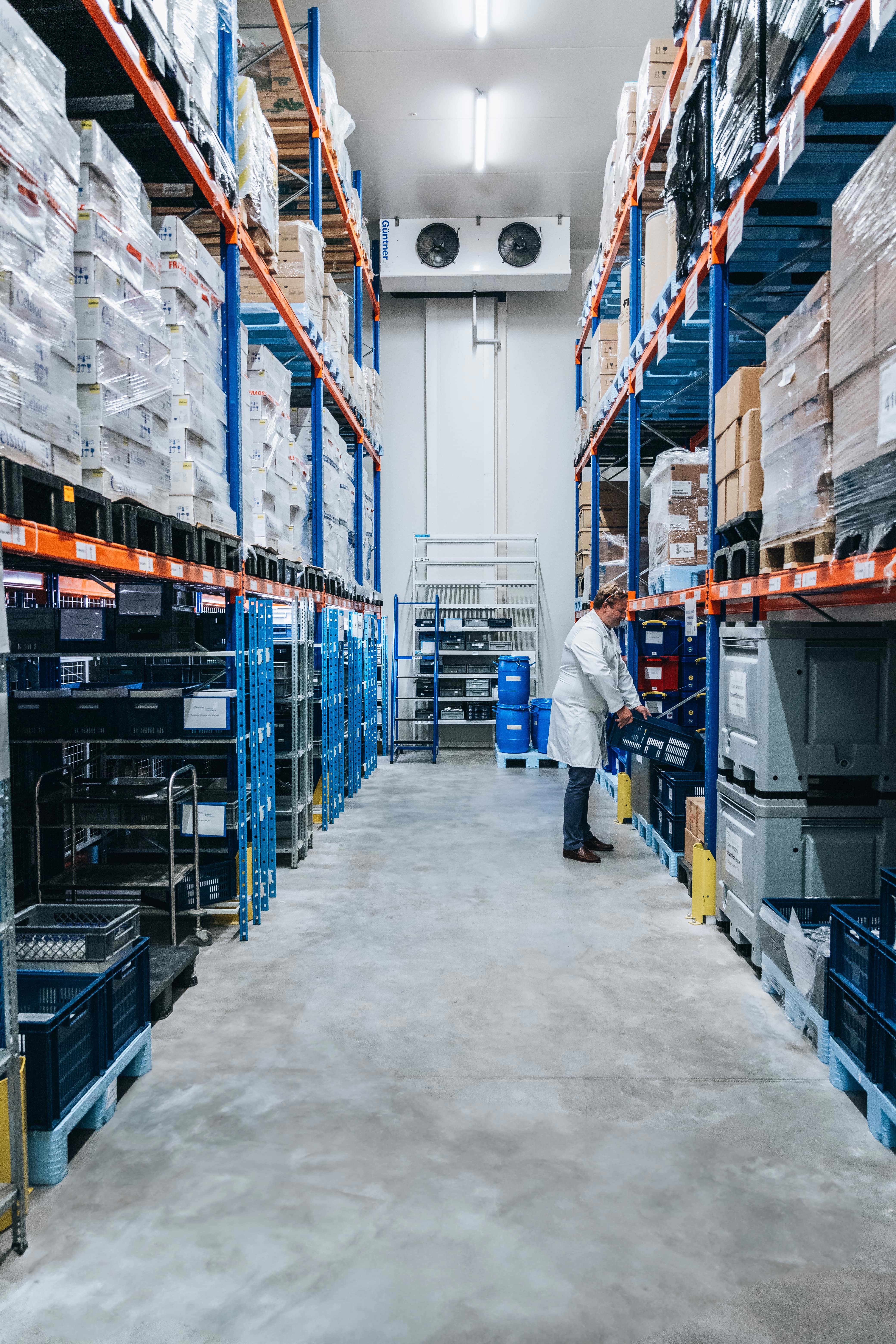 Zone de stockage d’Eurofins BioPharma Product Testing avec étagères métalliques bleues sur plusieurs niveaux, contenant des boîtes et des équipements. Un collaborateur en blouse blanche inspecte les étagères, illustrant la gestion rigoureuse des échantillons dans le cadre des études de stabilité selon les normes ICH et GMP.