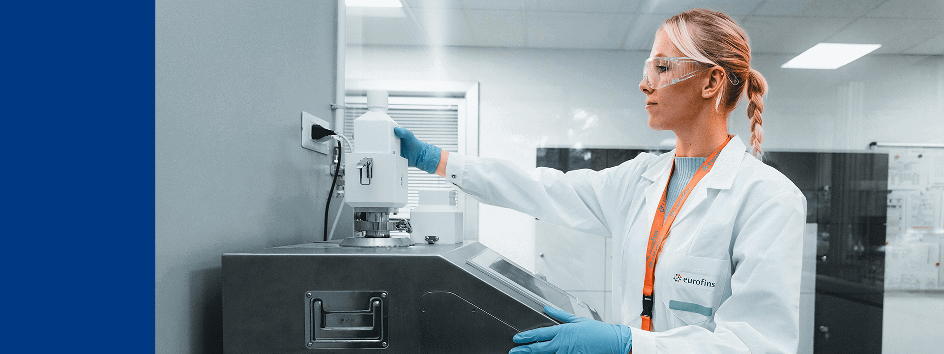 Un technicien d’Eurofins BioPharma Product Testing, équipé d’une blouse blanche, de gants bleus et de lunettes de protection, manipule un appareil analytique dans un laboratoire moderne. Le logo Eurofins est visible sur la blouse, et plusieurs équipements scientifiques sont présents en arrière-plan, illustrant les procédures de tests sur les contenants pharmaceutiques dans le cadre du contrôle qualité.