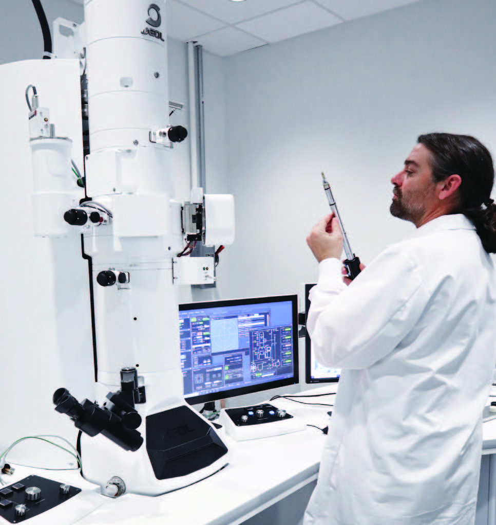 Scientifique Eurofins BioPharma Product Testing manipulant un porte-échantillon devant un microscope électronique JEOL dans un laboratoire pharmaceutique, avec des écrans affichant des données d’analyse avancées. Illustration de l’expertise en contrôle qualité, en microbiologie et en tests analytiques pour l’industrie biopharmaceutique.