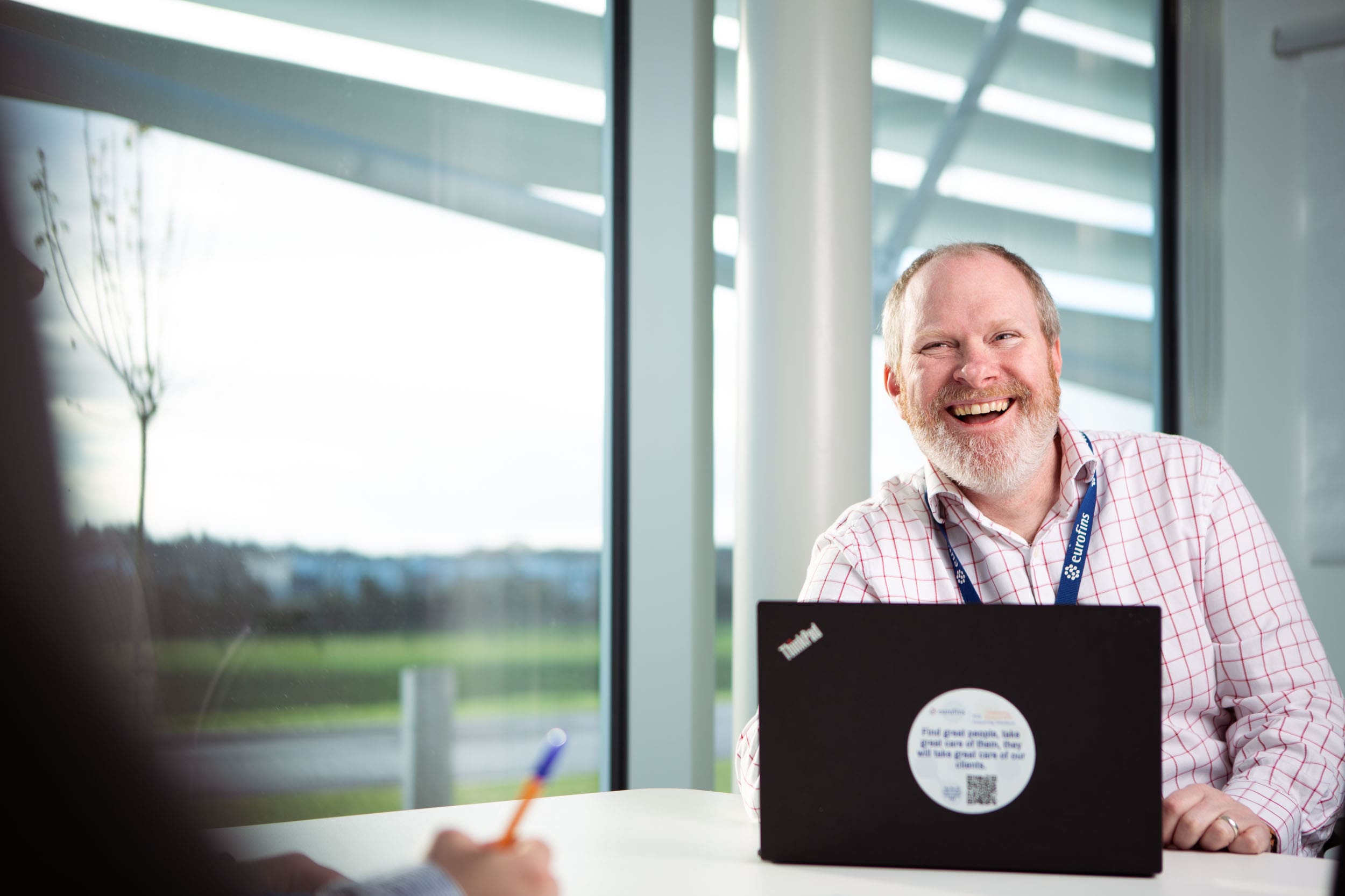 Un collaborateur d’Eurofins BioPharma Product Testing, vêtu d’une chemise à carreaux et portant un badge, est assis à une table devant un ordinateur portable Lenovo. Il sourit en regardant vers la gauche, tandis qu’une autre personne, partiellement visible au premier plan, prend des notes sur du papier. De grandes fenêtres donnent sur un extérieur verdoyant, illustrant un cadre professionnel ouvert et collaboratif, dédié aux études sur les matériaux en verre utilisés dans les emballages pharmaceutiques.
