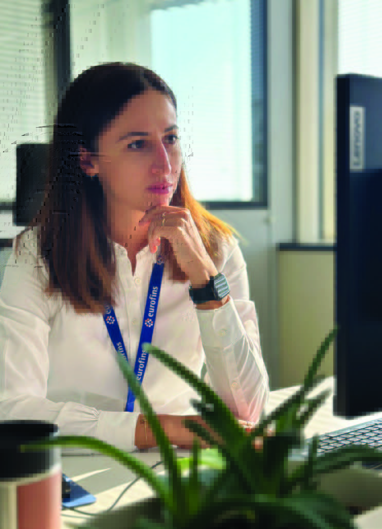 Collaborateur Eurofins Biopharma Product Testing assis à un bureau, portant un badge Eurofins autour du cou et travaillant sur un ordinateur Lenovo dans un bureau lumineux. Au premier plan, une plante verte et une tasse sont visibles sur la table.