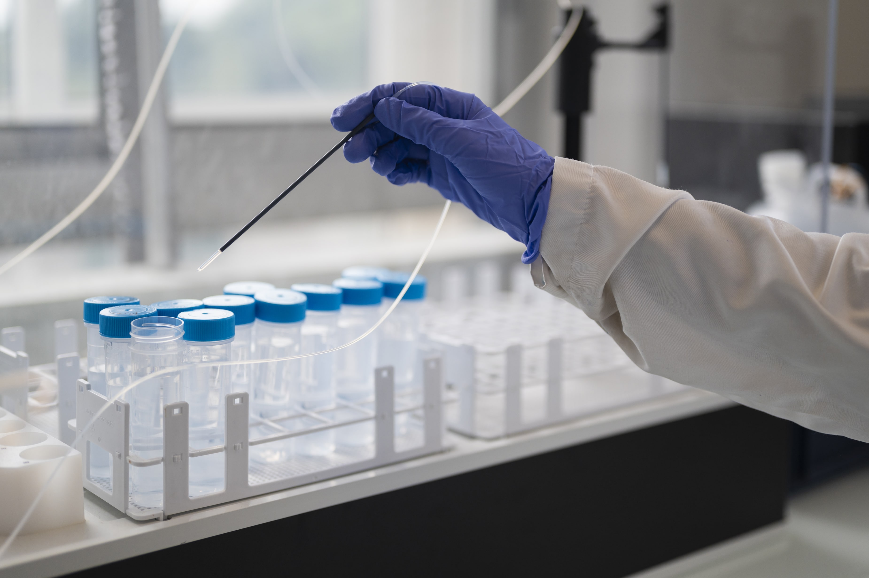 Un technicien en laboratoire, portant une blouse et des gants bleus, manipule une tige fine au-dessus d’un rack contenant plusieurs tubes à essai avec bouchons bleus. Cette scène illustre les activités d’analyses et de préparation d’échantillons réalisées par Eurofins BioPharma Product Testing.