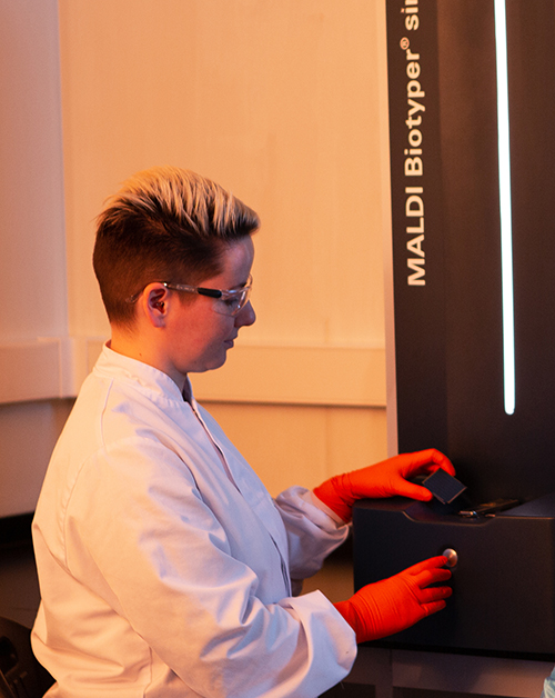 Scientifique en blouse de laboratoire et gants orange manipulant un échantillon sur un appareil MALDI Biotyper® dans un environnement analytique, illustrant les méthodes avancées d’identification microbienne utilisées chez Eurofins BioPharma Product Testing.