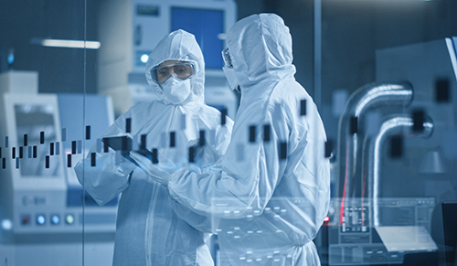 Techniciens en combinaison de protection intégrale dans un laboratoire pharmaceutique haute technologie, examinant une tablette électronique au cœur d’un environnement stérile. Cette image illustre les opérations critiques de la logistique des essais cliniques chez Eurofins Biopharma Product Testing France, incluant le conditionnement, la traçabilité et le contrôle qualité des médicaments expérimentaux selon les normes GMP et les exigences réglementaires internationales.