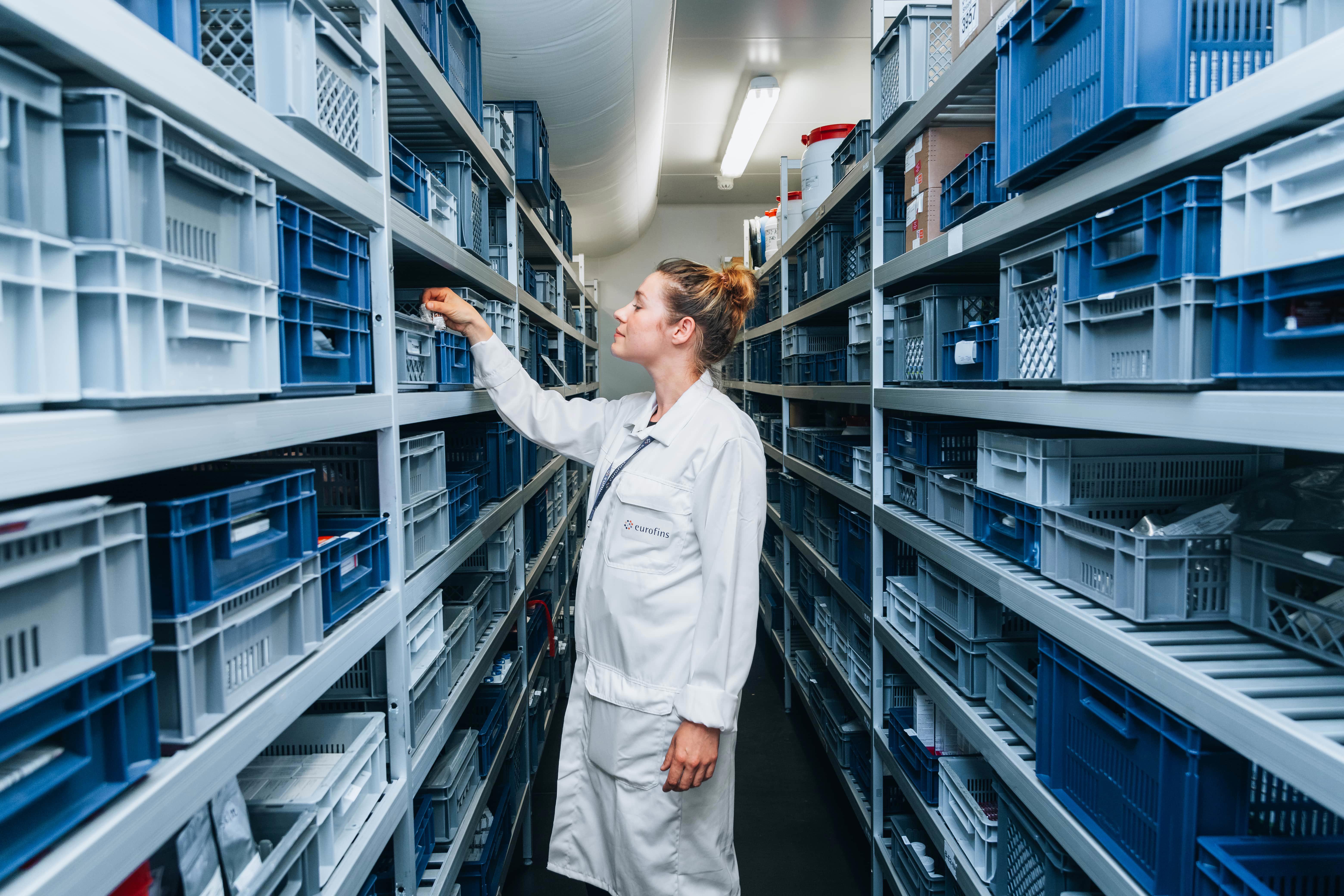 Employé en blouse blanche chez Eurofins BioPharma Product Testing dans un entrepôt de stockage organisé, manipulant des bacs en plastique sur des étagères métalliques. L’image illustre les pratiques de gestion et de traçabilité dans le cadre des essais de stabilité des produits biologiques et biotechnologiques, conformément aux lignes directrices ICH.