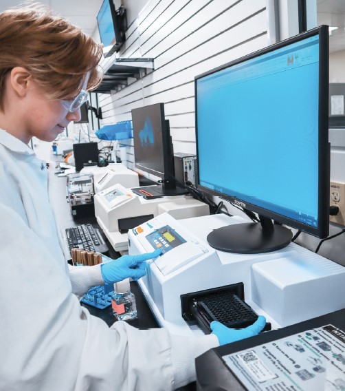 Personne en blouse blanche, lunettes de protection et gants, manipulant une microplaque dans un laboratoire équipé d’appareils automatisés. Elle utilise un poste informatique affichant une interface bleue, dans un environnement dédié aux analyses scientifiques. Cette scène illustre les activités de recherche et de tests menées par Eurofins BioPharma Product Testing France.