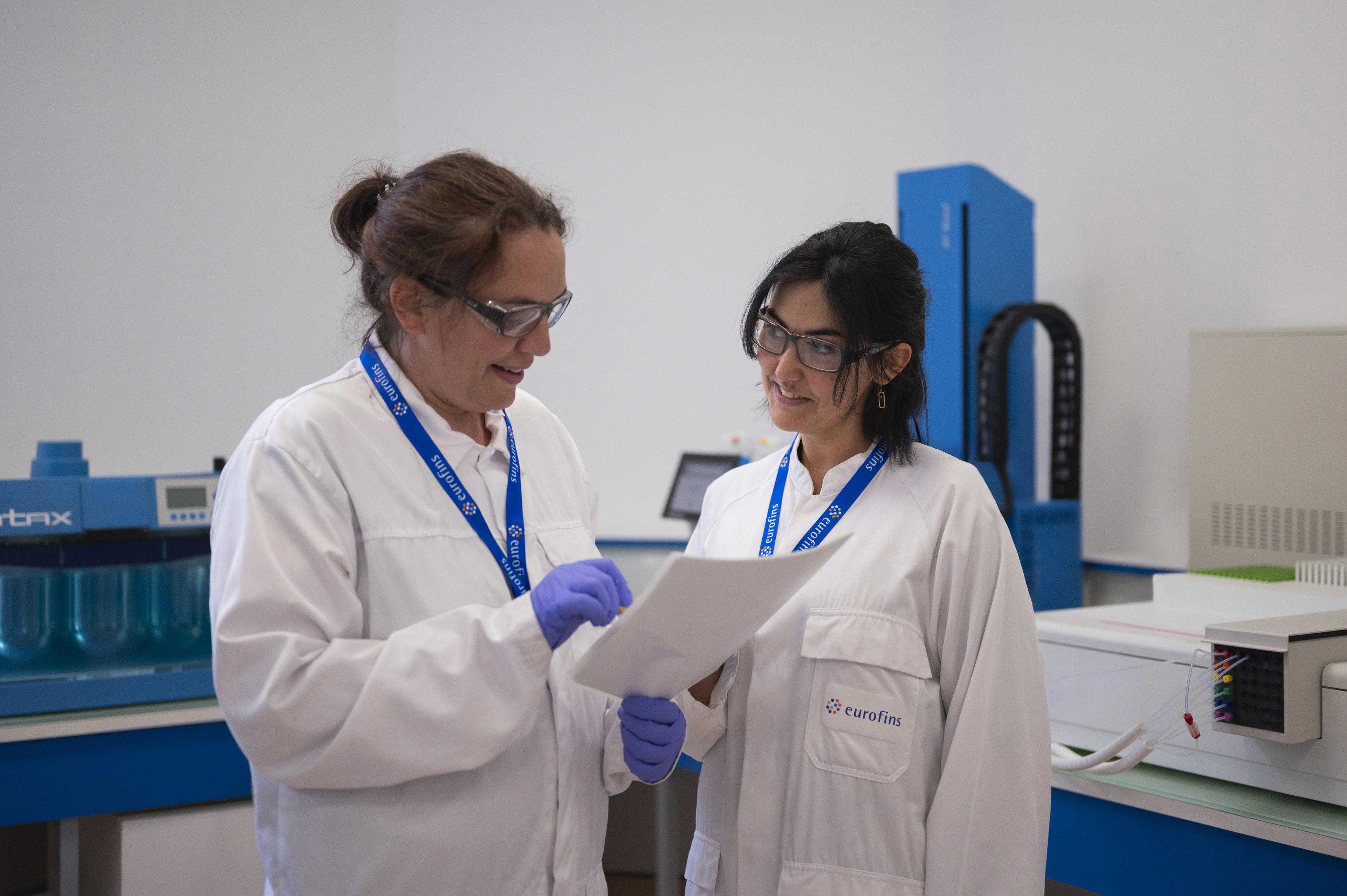 Deux collaborateurs en blouse blanche siglée Eurofins et lunettes de protection, discutant autour d’un document technique dans un laboratoire équipé d’instruments analytiques, dont un appareil "Vortex". L’image illustre une phase de collaboration scientifique dans le cadre des études de dissolution menées par Eurofins BioPharma Product Testing.