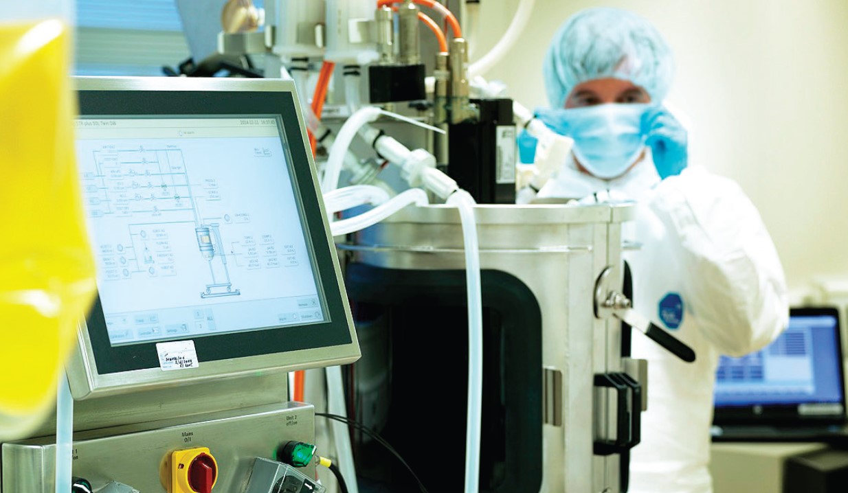 Équipement de bioproduction dans un laboratoire Eurofins BioPharma Product Testing, comprenant un bioréacteur en acier inoxydable relié à des tuyaux et capteurs, ainsi qu’un écran de contrôle affichant un schéma technique du procédé. À l’arrière-plan, un poste informatique est visible, illustrant un environnement de production pharmaceutique conforme aux standards de qualité.