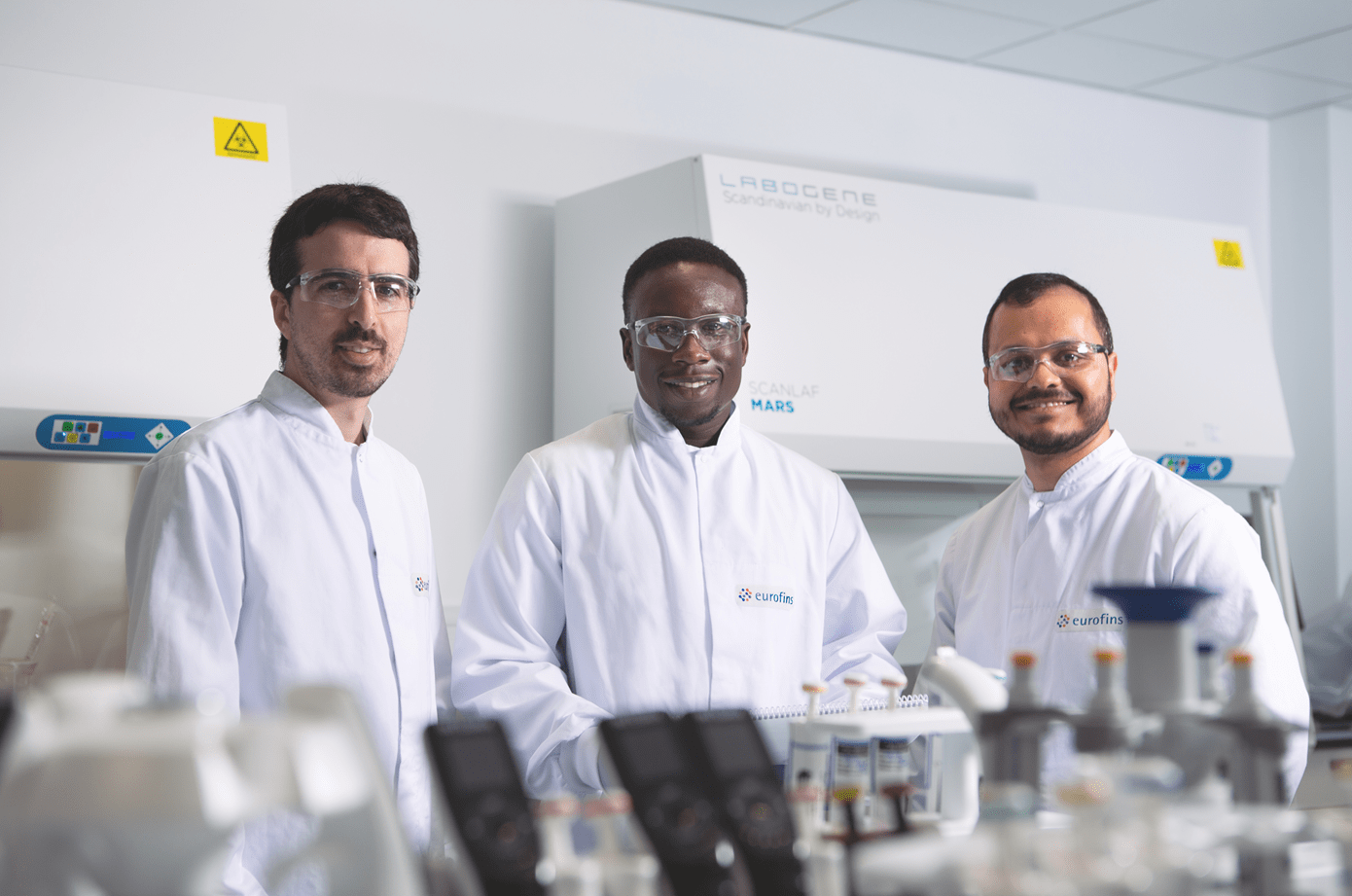 Trois techniciens de laboratoire chez Eurofins BioPharma Product Testing portant des blouses blanches, positionnés devant un équipement de sécurité biologique de type hotte. Sur la paillasse, plusieurs instruments et flacons sont visibles, illustrant un environnement conforme aux normes GMP pour la réalisation d’analyses biopharmaceutiques.