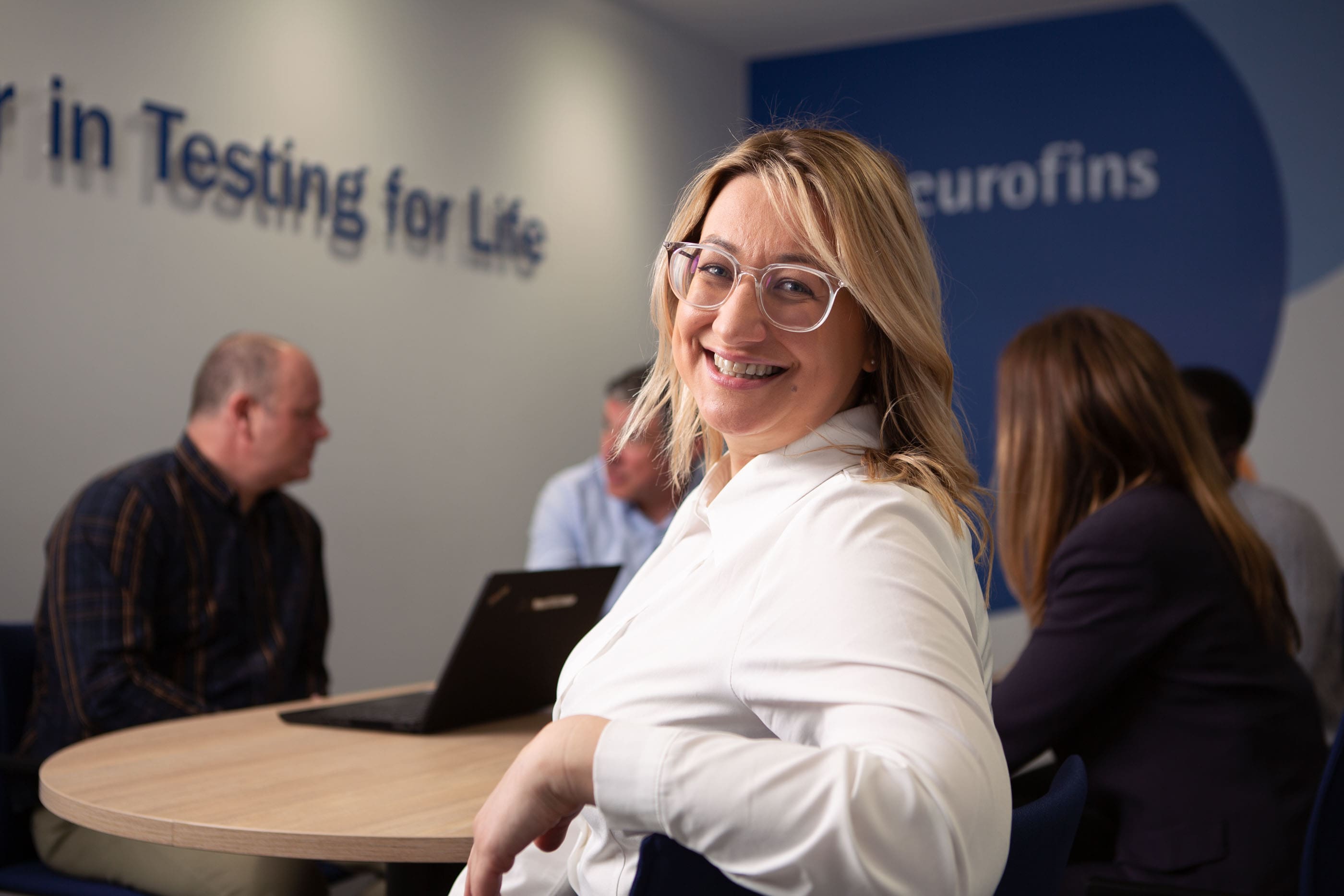 Réunion d’experts chez Eurofins BioPharma Product Testing dans un espace collaboratif dédié aux services d’analyse pharmaceutique. Un collaborateur en chemise blanche sourit au premier plan, tandis que d’autres professionnels échangent autour d’un ordinateur portable. Le slogan 'Testing for Life' et le logo Eurofins visibles en arrière-plan renforcent l’engagement de l’entreprise dans la consultation scientifique, le développement analytique et le support technique pour l’industrie des sciences de la vie.