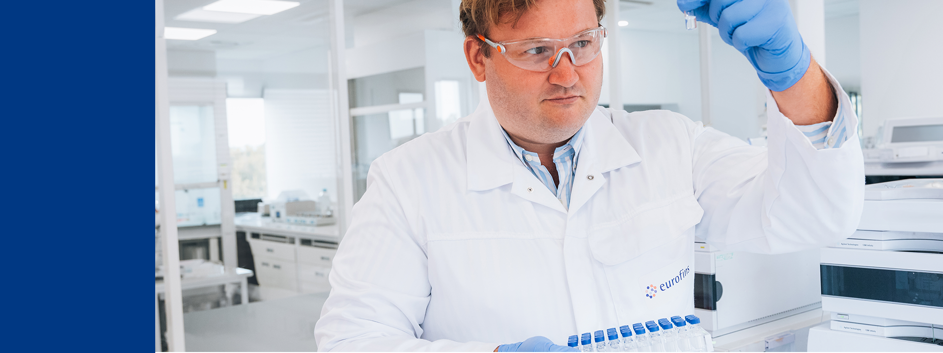 Un technicien de laboratoire en blouse blanche, gants bleus et lunettes de protection examine un échantillon dans un environnement pharmaceutique moderne. Il tient un rack de tubes à essai, illustrant les étapes de contrôle qualité réalisées par Eurofins Biopharma Product Testing dans le cadre de la libération complète des produits pour l’Union européenne, avec certification QP conforme aux exigences réglementaires