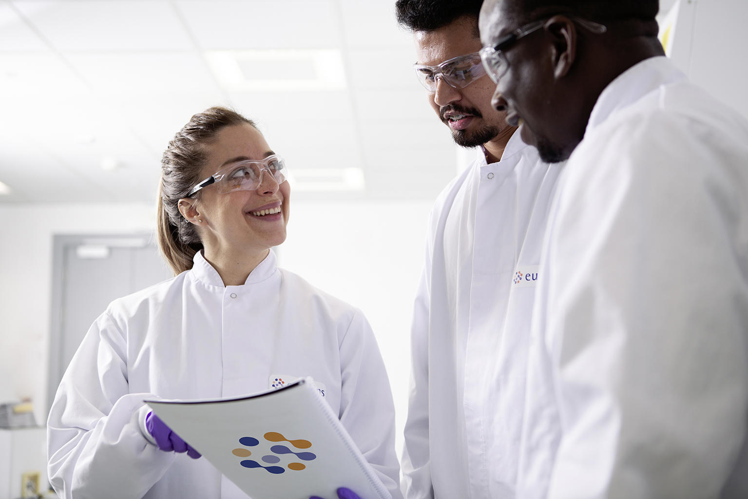 Professionnels en blouses de laboratoire Eurofins BioPharma Product Testing discutant dans un environnement scientifique moderne, tenant un document avec le logo Eurofins. L’image illustre la collaboration technique et la rigueur des procédures dans le cadre du contrôle qualité et des analyses pharmaceutiques conformes aux normes GMP et ISO.