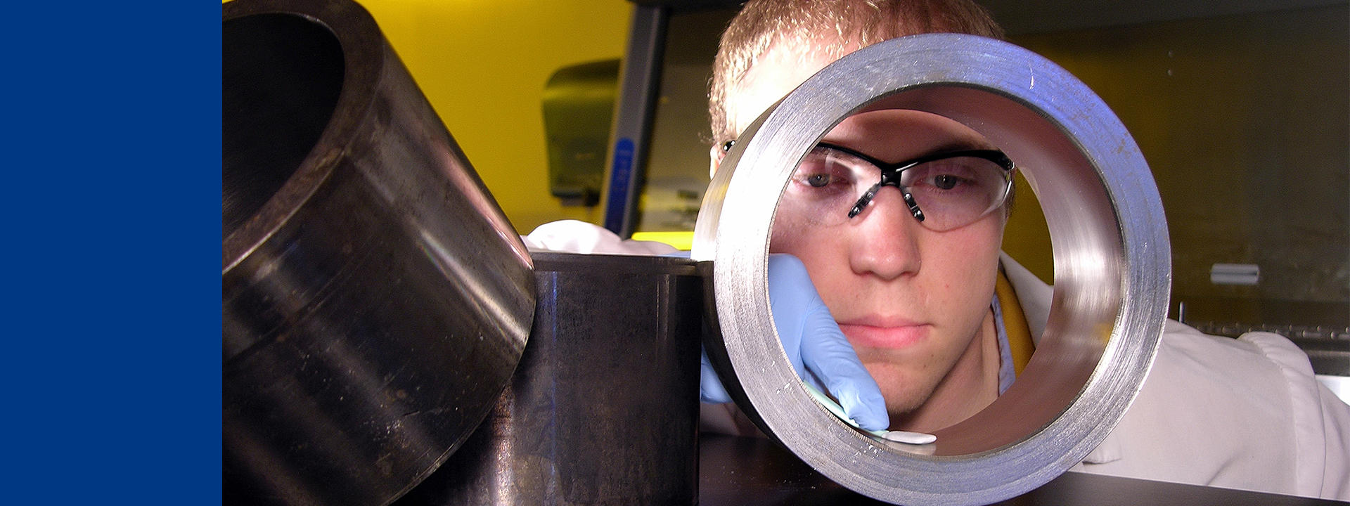 Un technicien de laboratoire en blouse blanche, lunettes de sécurité et gants examine un composant cylindrique métallique dans un environnement scientifique. Cette inspection illustre les services de validation des installations et des procédés proposés par Eurofins Biopharma Product Testing, garantissant la conformité aux normes BPF dans le secteur pharmaceutique.