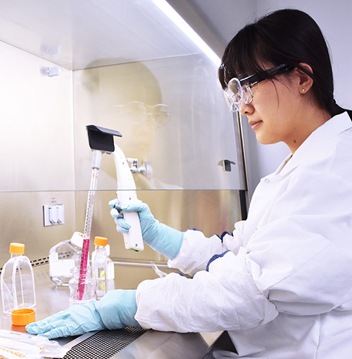 Scientifique en blouse de laboratoire, lunettes de protection et gants bleus manipulant des liquides à l’aide d’une pipette dans un environnement stérile. Des flacons, bouteilles et un cylindre gradué rose sont disposés sur la paillasse. Cette scène illustre les activités de laboratoire menées par Eurofins BioPharma Product Testing dans le cadre de ses analyses biologiques et chimiques de haute précision.