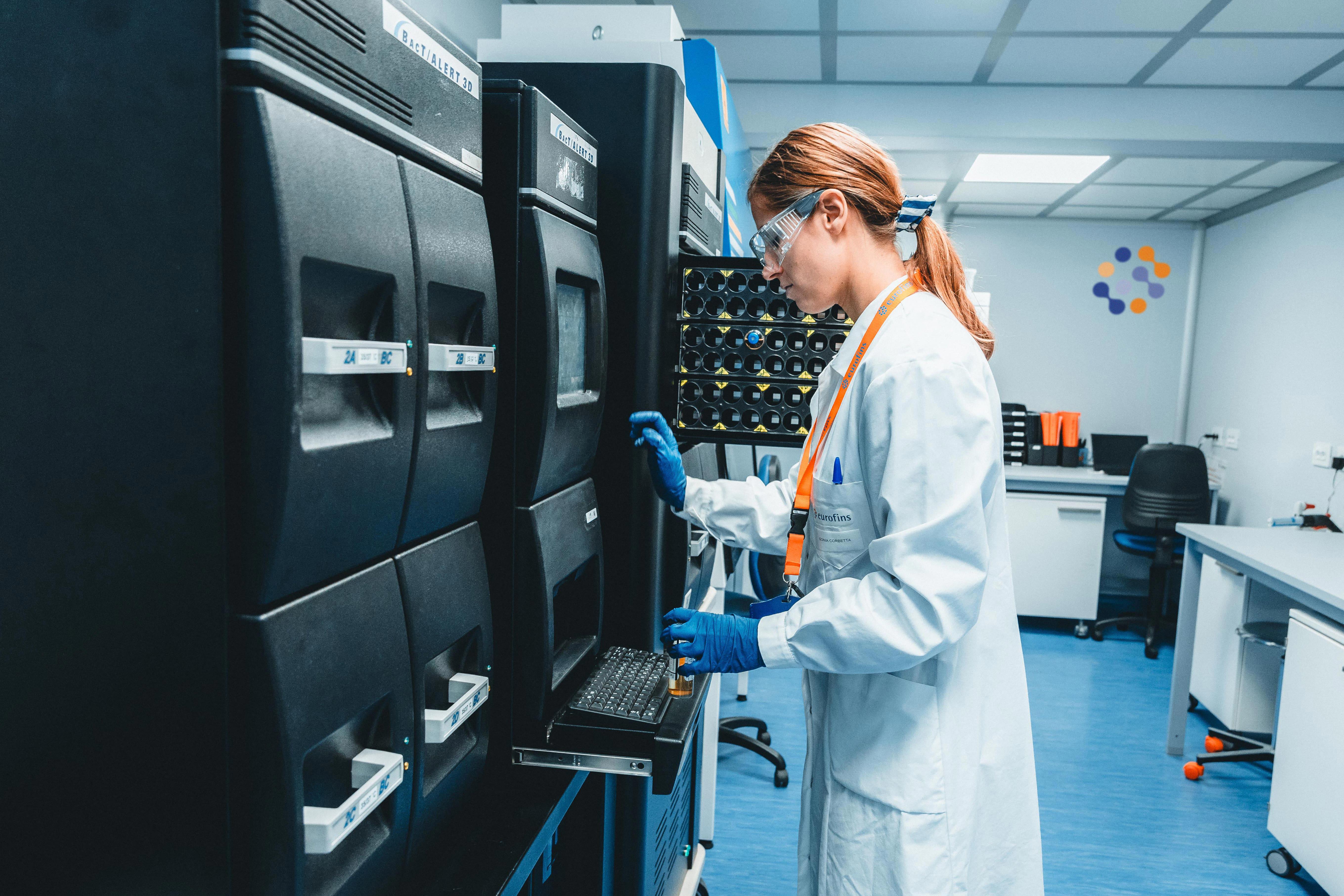 Technicien de laboratoire Eurofins Biopharma Product Testing portant une blouse blanche et des gants bleus, manipulant un système automatisé BACT/ALERT pour la détection microbiologique. L’image montre un environnement de laboratoire moderne avec plusieurs instruments alignés et un plan de travail équipé.