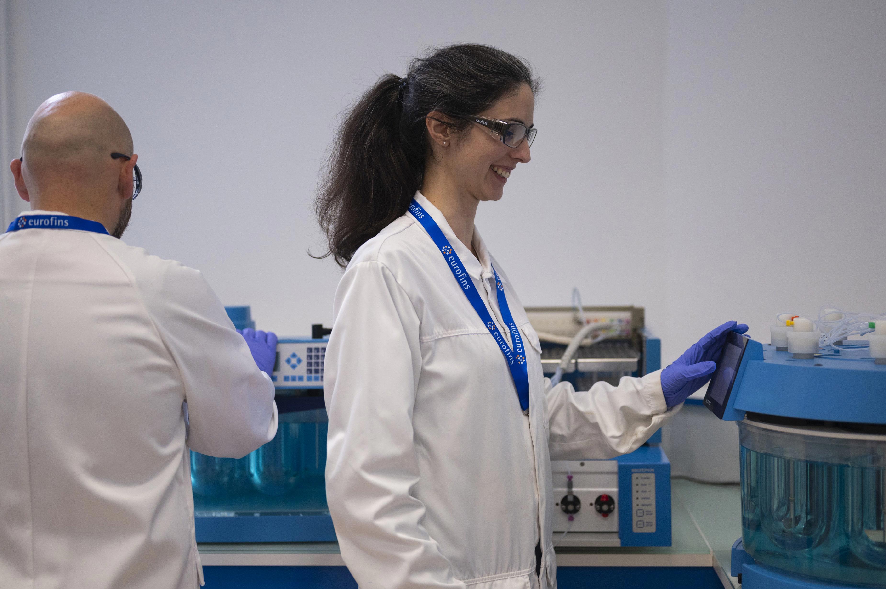 Deux collaborateurs en blouse blanche, gants bleus et lunettes de protection, travaillant dans un laboratoire équipé d’instruments analytiques. L’un interagit avec un appareil doté d’un écran digital, tandis que l’autre manipule du matériel en arrière-plan. Les deux portent des badges Eurofins. L’image illustre les opérations de test de dissolution réalisées par Eurofins BioPharma Product Testing dans un environnement scientifique conforme aux normes pharmaceutiques.