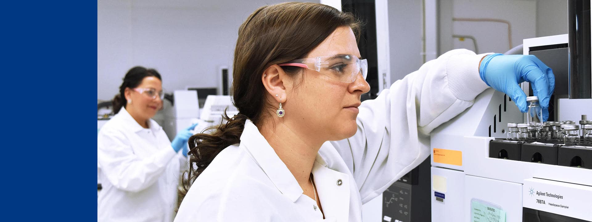 Technicien de laboratoire chez Eurofins BioPharma Product Testing manipulant des flacons sur un système analytique de chromatographie en phase gazeuse. La personne porte une blouse blanche et des gants de protection bleus, illustrant les procédures de sécurité appliquées dans un environnement de laboratoire conforme aux normes GMP. À l’arrière-plan, un autre poste de travail avec des équipements analytiques est visible, soulignant la précision et la rigueur des analyses réalisées par Eurofins.
