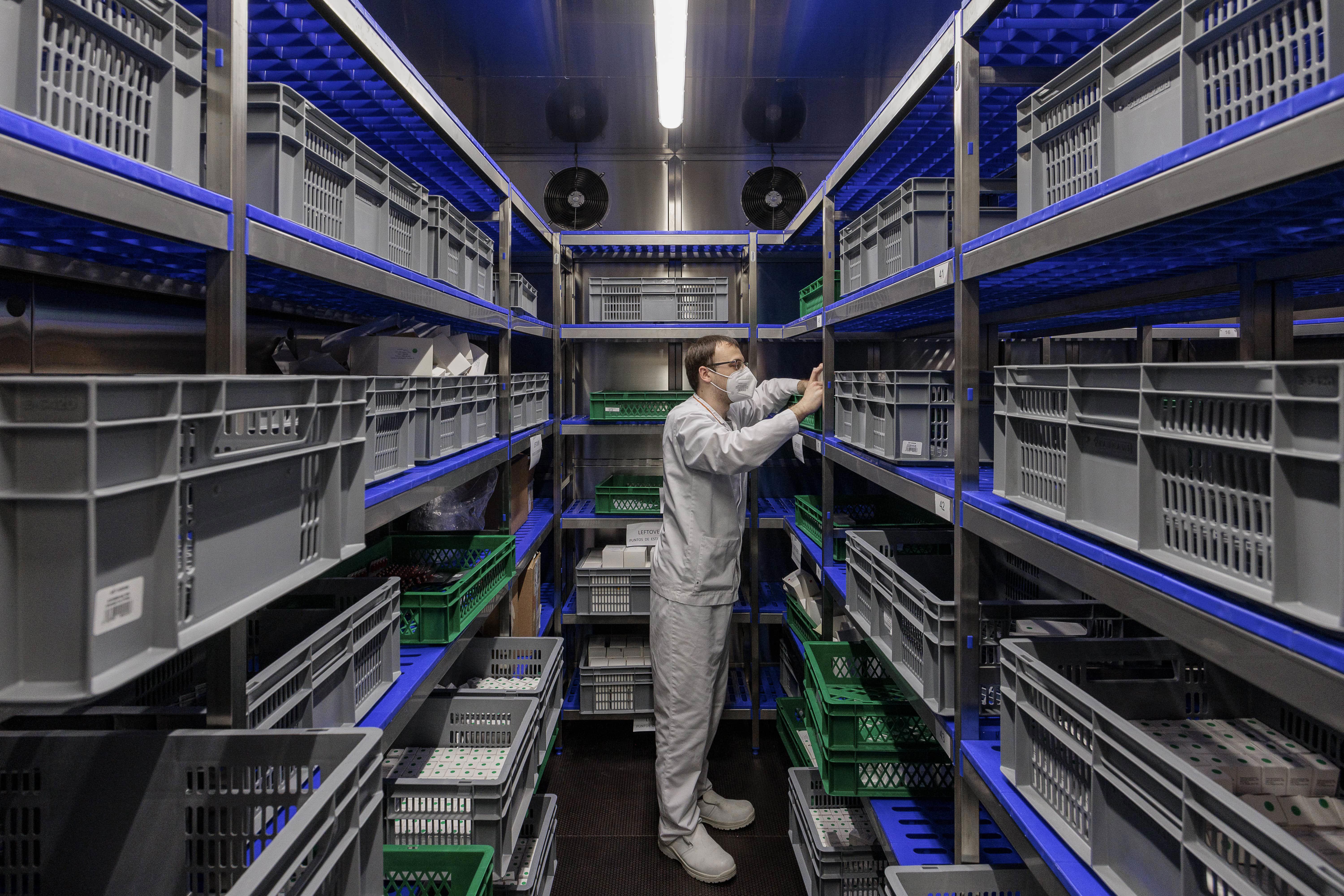 Technicien de laboratoire Eurofins BioPharma Product Testing en blouse blanche, debout dans une salle de stockage équipée d’étagères métalliques contenant des bacs en plastique gris et vert. Il inspecte ou manipule un objet sur une étagère intermédiaire, sous un éclairage fluorescent.