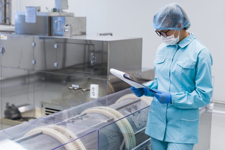 Technicienne de laboratoire en tenue de laboratoire stérile (charlotte, masque, lunettes de protection) examinant un document à côté d’un équipement industriel en acier inoxydable. Illustration utilisée par Eurofins BioPharma Product Testing pour représenter les essais sur les matières premières dans un environnement contrôlé conforme aux normes pharmaceutiques.