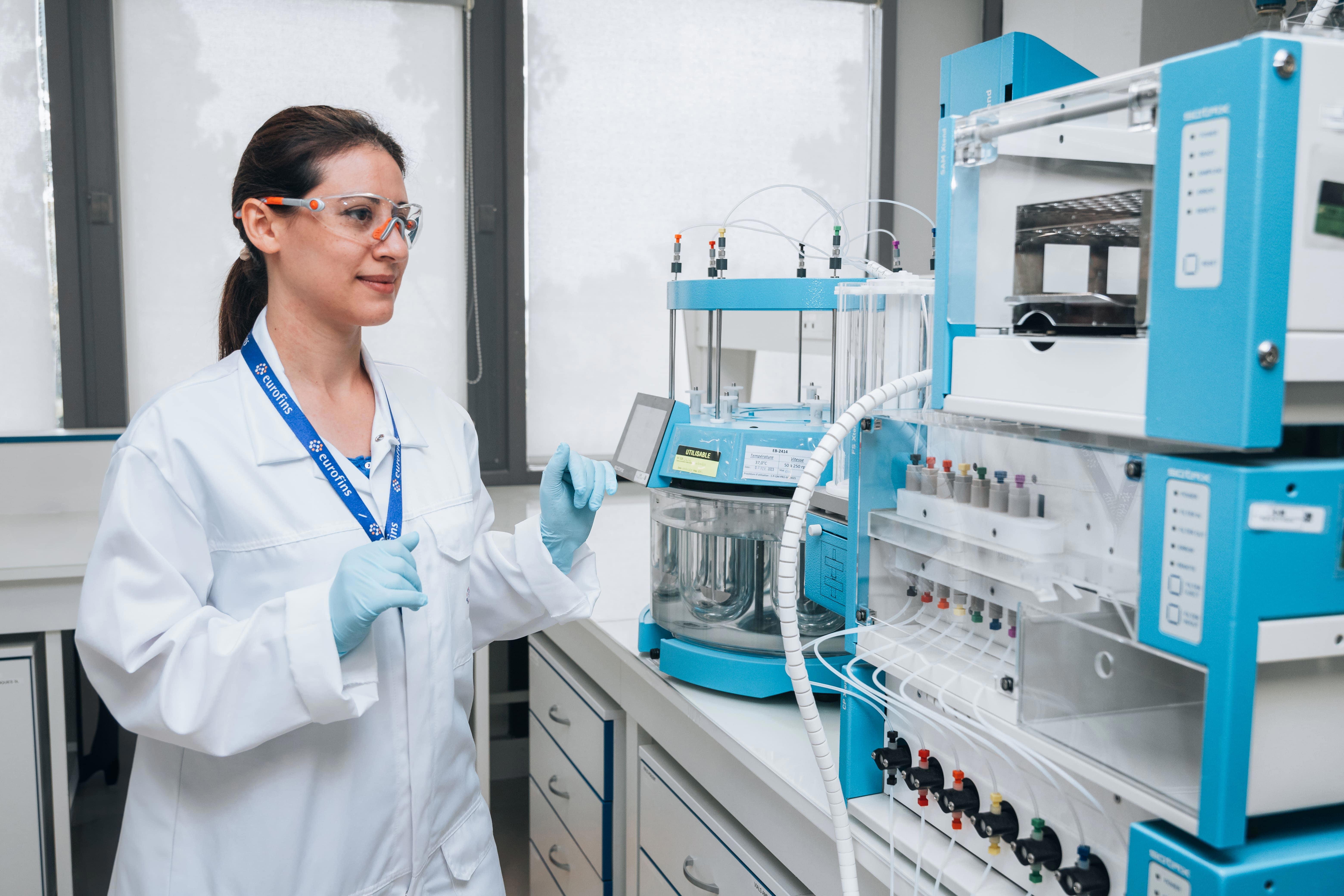 Personnel de laboratoire équipé d’une blouse blanche, de lunettes de protection et d’un badge, positionné à côté d’un dispositif analytique complexe composé de tubes et récipients interconnectés. Cette scène illustre les environnements techniques et les équipements de pointe utilisés par Eurofins BioPharma Product Testing dans le cadre de ses essais comparatifs et de ses analyses de produits pharmaceutiques.