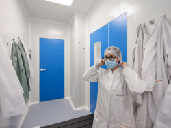 Technicien d’Eurofins BioPharma Product Testing dans une salle blanche, ajustant son masque chirurgical dans un environnement stérile conforme aux normes GMP. Vêtu d’une blouse blanche et d’un bonnet, il se prépare à intervenir dans une zone de contrôle microbiologique dédiée à la détection d’organismes spécifiques. Des équipements de protection individuelle sont suspendus au mur, illustrant les protocoles stricts d’hygiène et de biosécurité appliqués dans les laboratoires pharmaceutiques.