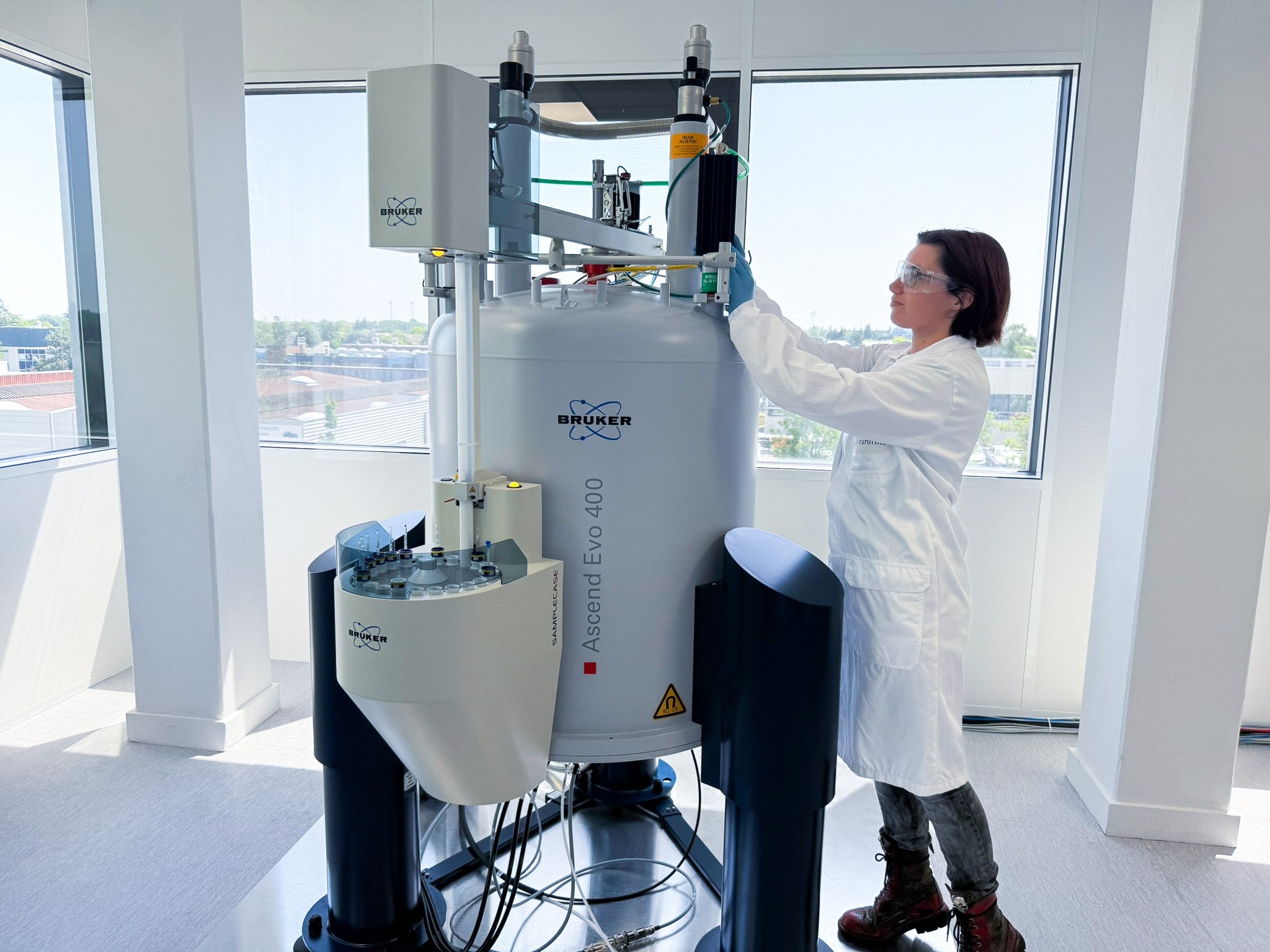Scientifique en blouse Eurofins, équipé de lunettes de protection, manipulant un spectromètre RMN Bruker Ascend Evo 400 MHz dans un laboratoire lumineux. Cette technologie de résonance magnétique nucléaire permet à Eurofins BioPharma Product Testing de caractériser avec précision la structure moléculaire des substances pharmaceutiques dans le cadre de leurs analyses avancées.