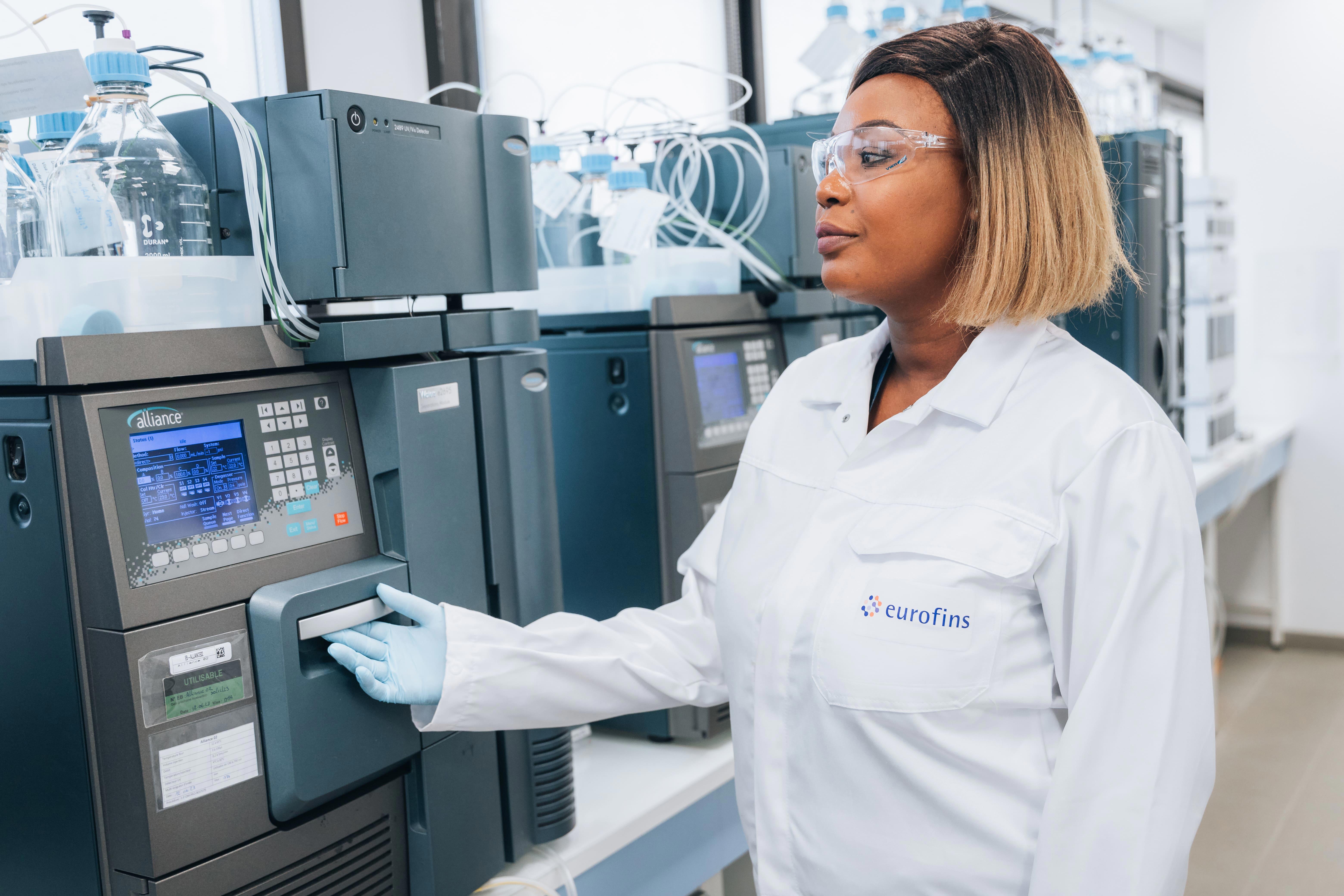 Scientifique en blouse blanche Eurofins, équipé de lunettes de protection et de gants, opérant un système HPLC Alliance dans un laboratoire. L’environnement technique comprend divers équipements analytiques et flacons reliés par des tubes, illustrant les processus de validation d’études et d’interprétation statistique des données menés par Eurofins BioPharma Product Testing.