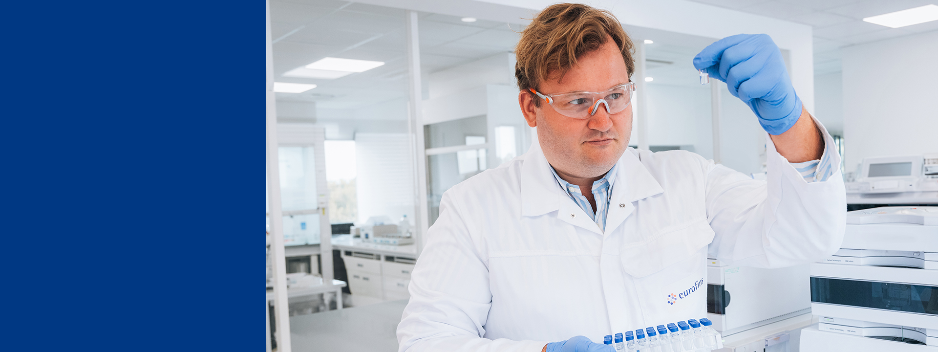 Technicien Eurofins BioPharma Product Testing manipulant un flacon d’échantillon liquide dans un laboratoire certifié BPF, tenant un rack de vials pour analyses physicochimiques (pH, pureté, composition) et microbiologiques (charge microbienne, stérilité) dans le cadre d’études de stabilité pharmaceutique selon les directives ICH, assurant contrôle qualité, validation des conditions de stockage et détermination de la durée de conservation des médicaments.