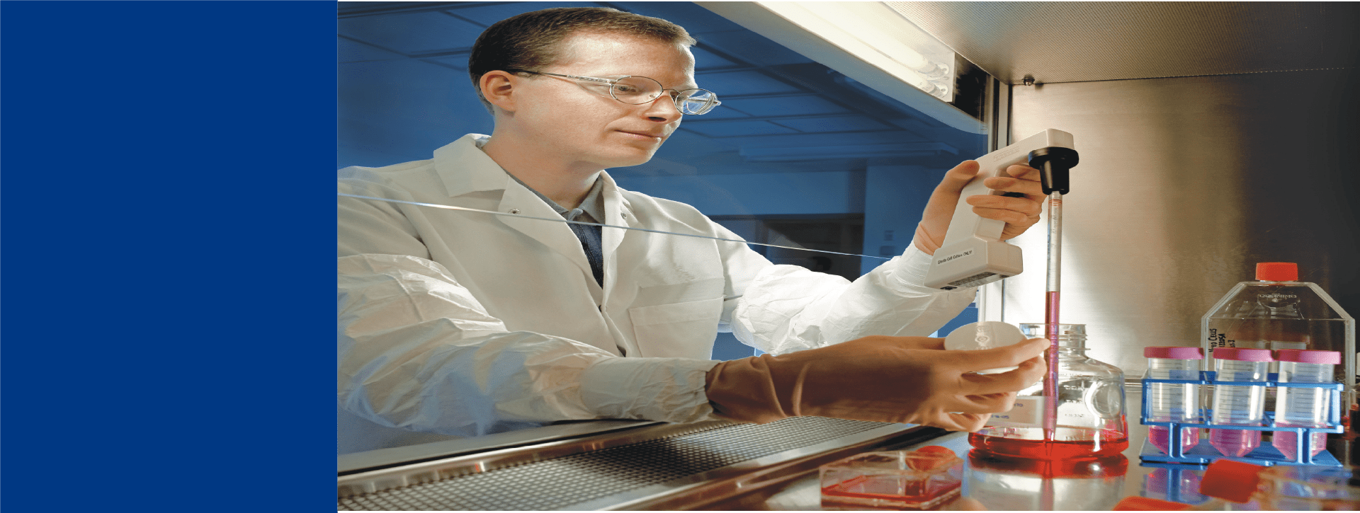 Technicien de laboratoire chez Eurofins BioPharma Product Testing manipulant une pipette électronique sous une hotte à flux laminaire. Sur la paillasse, plusieurs flacons et boîtes de culture contenant un milieu rouge sont disposés, illustrant les procédures stériles appliquées pour les tests de dépistage des mycoplasmes conformément aux normes GMP.